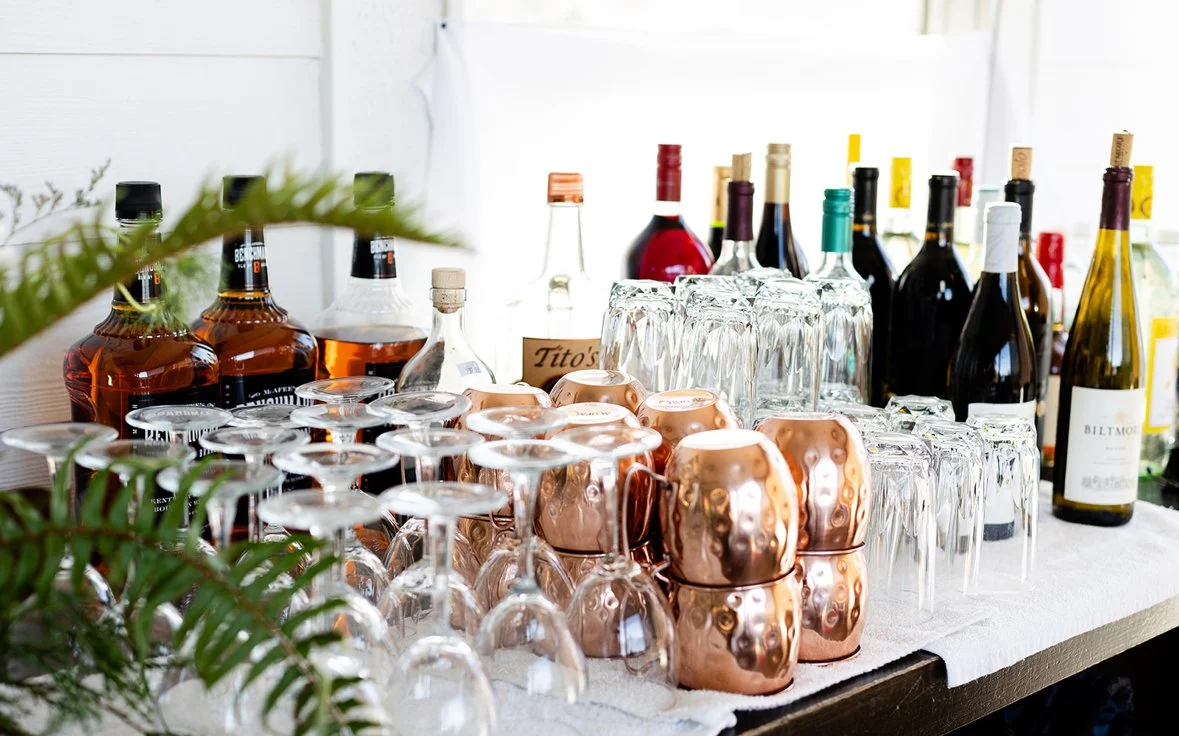 An assortment of alcohol bottles, glassware, and copper mugs on a table, with some greenery in the foreground.