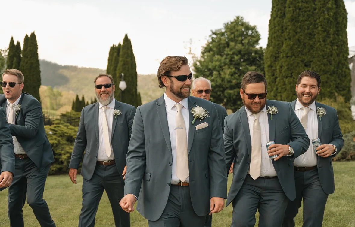 Groom and groomsmen in matching gray suits and sunglasses smiling outdoors during a wedding celebration.