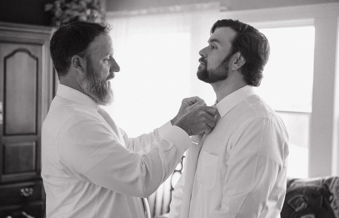 An older man helps a young man adjust his tie in a bedroom with a wooden wardrobe and window with curtains.