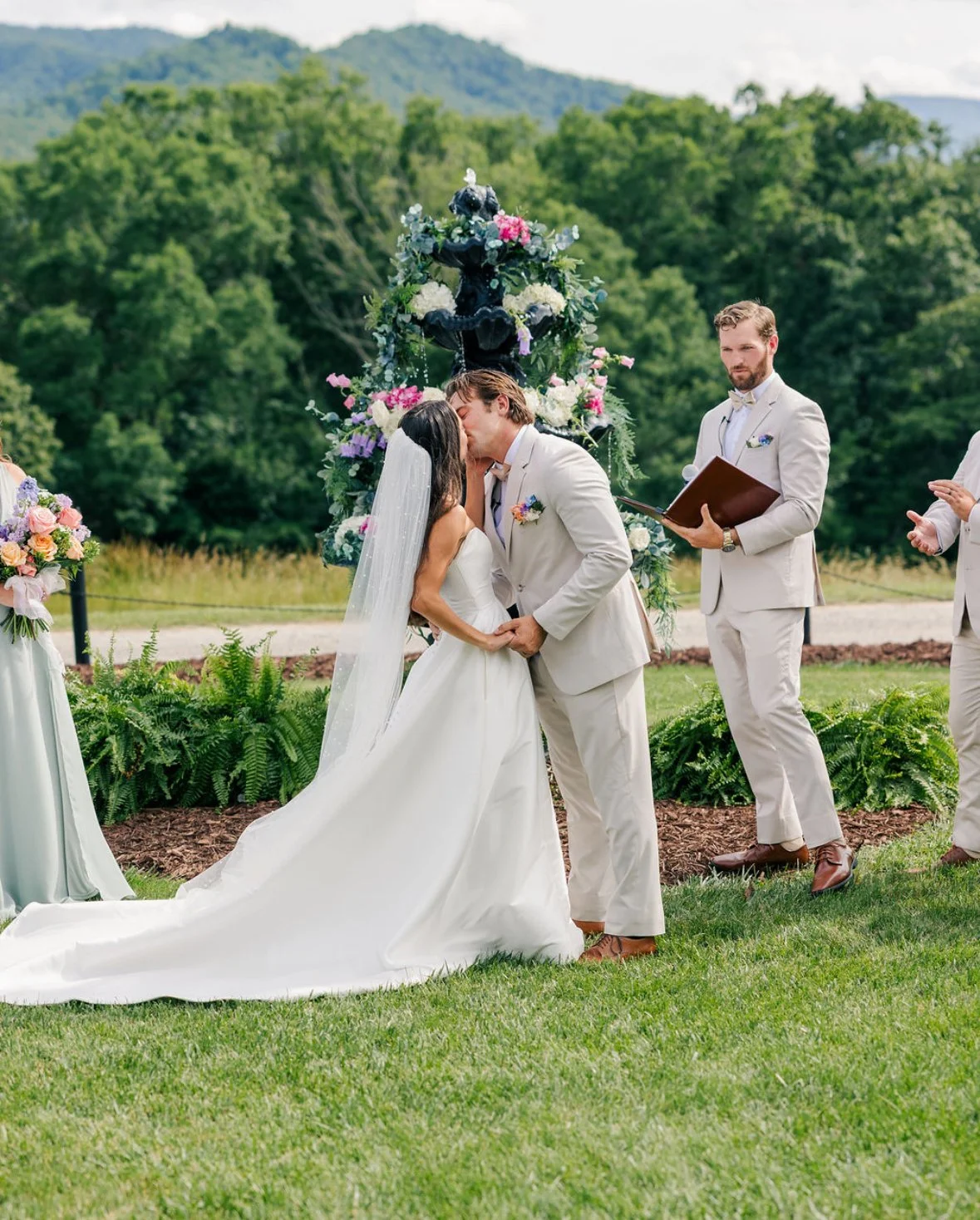 A bride and groom share a kiss during their outdoor wedding ceremony, with a colorful floral backdrop and officiant nearby.