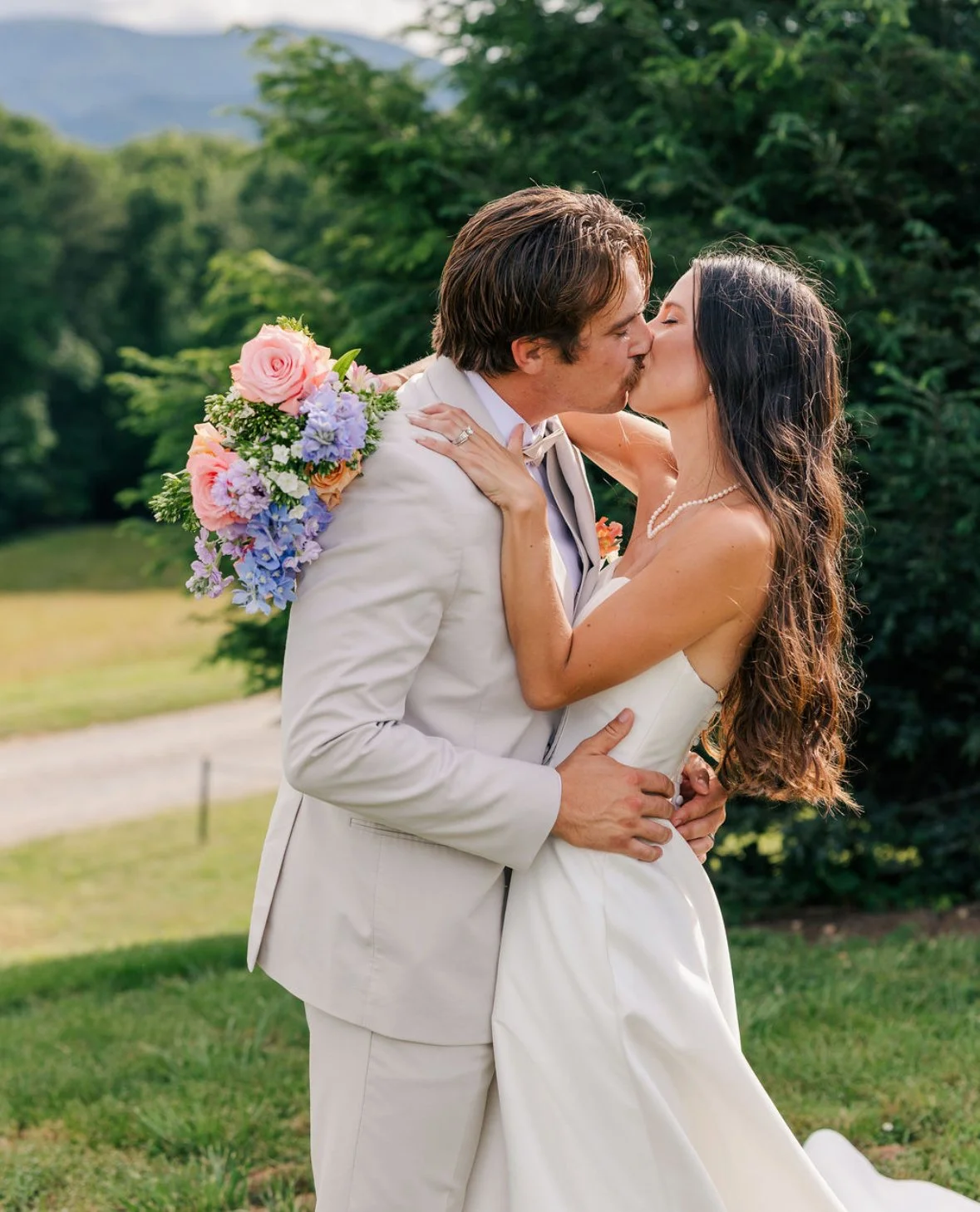 A bride and groom kissing outdoors on a sunny day, the bride holding a bouquet of pink, purple, and white flowers, with greenery and trees in the background.