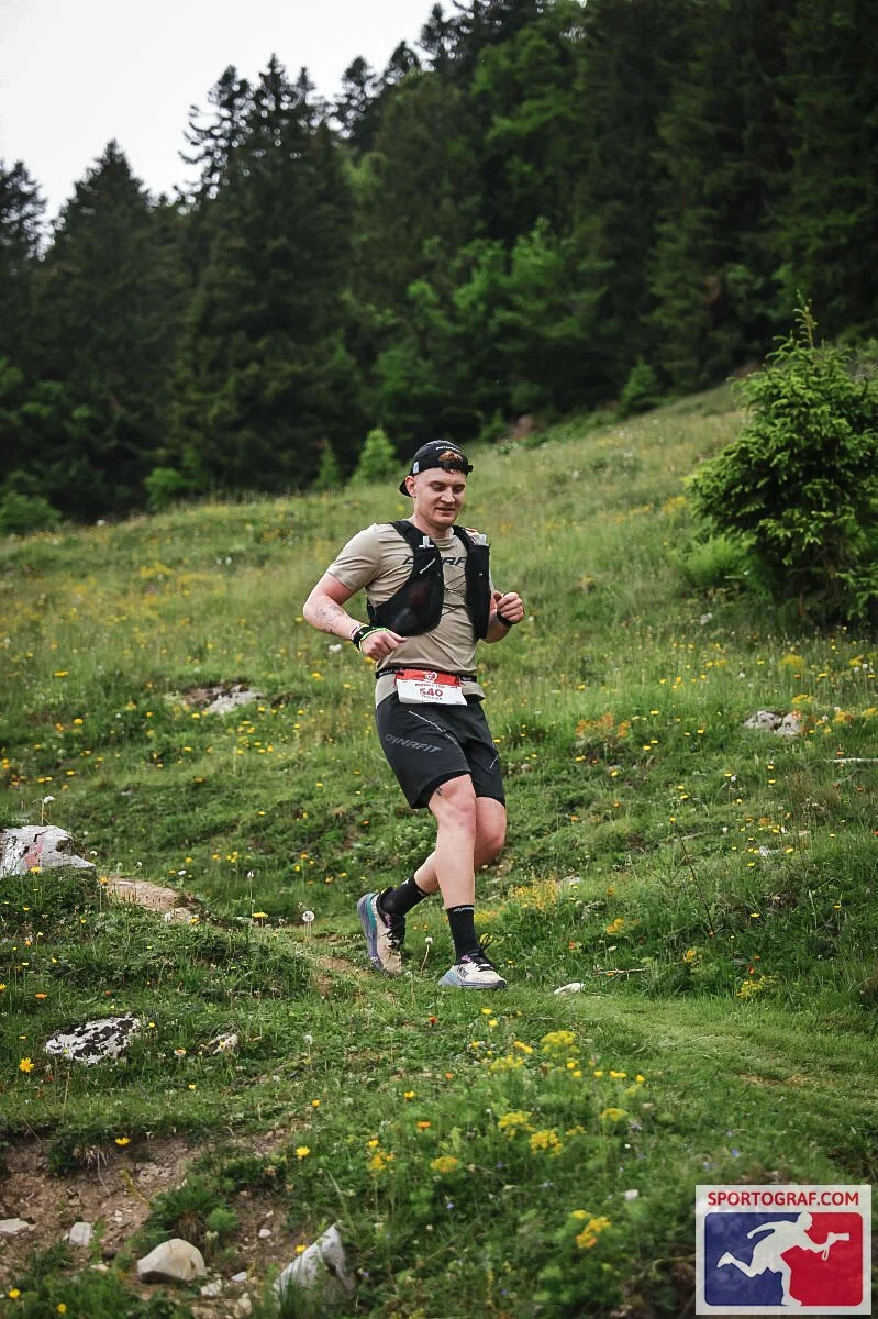 Ein Mann läuft auf einem bewaldeten Hang, trägt eine Neongelbe T-Shirt, schwarze Shorts, einen Rucksack und eine Mütze. Es ist ein wildes, grünes Gelände mit Blumen und Steinen.