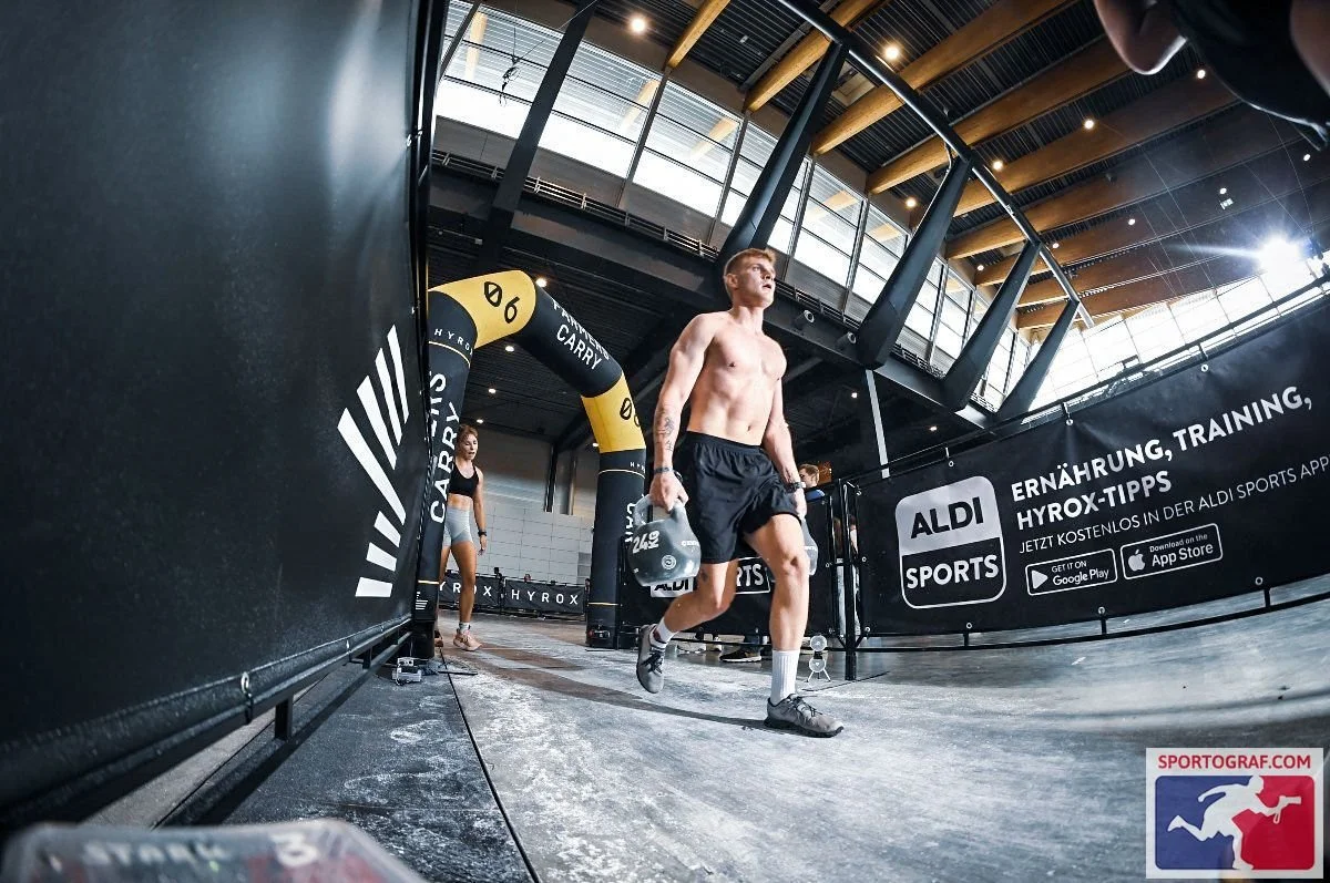 Ein Mann trainiert in einer Halle, trägt schwarze Shorts, hebt Hanteln und ist nackt, mit anderen Personen im Hintergrund. Hyrox Wettkampf