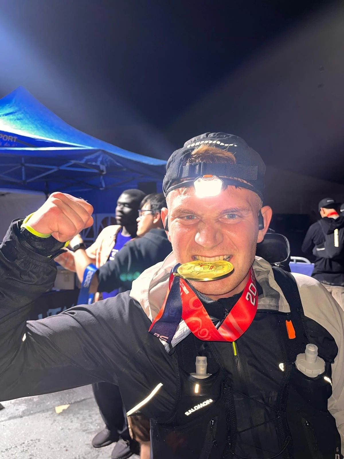 Ein Mann trägt eine Medaille im Mund, ein Finisher-Medaillenband um den Hals und eine Stirnlampe auf dem Kopf. Er ist bei einer nächtlichen Laufveranstaltung und zeigt einen heldenhaften Gesichtsausdruck. Ein Ultramarathon, Mozart100.
