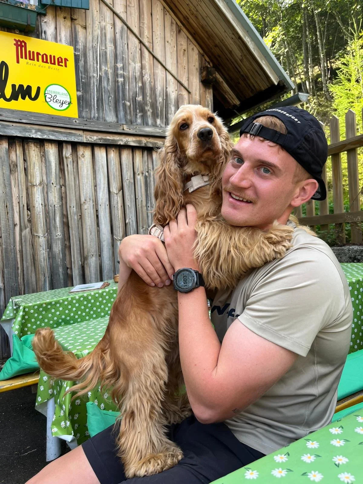 Ein lachender Mann hält einen goldene Cocker Spaniel-Hund in seinen Armen. Sie sitzen auf einer grünen, mit weißen Blumen bedruckten Tischdecke im Freien, hinter einer Holzwand. Der Mann trägt eine schwarze Kappe, ein beige T-Shirt und eine Sportuhr.