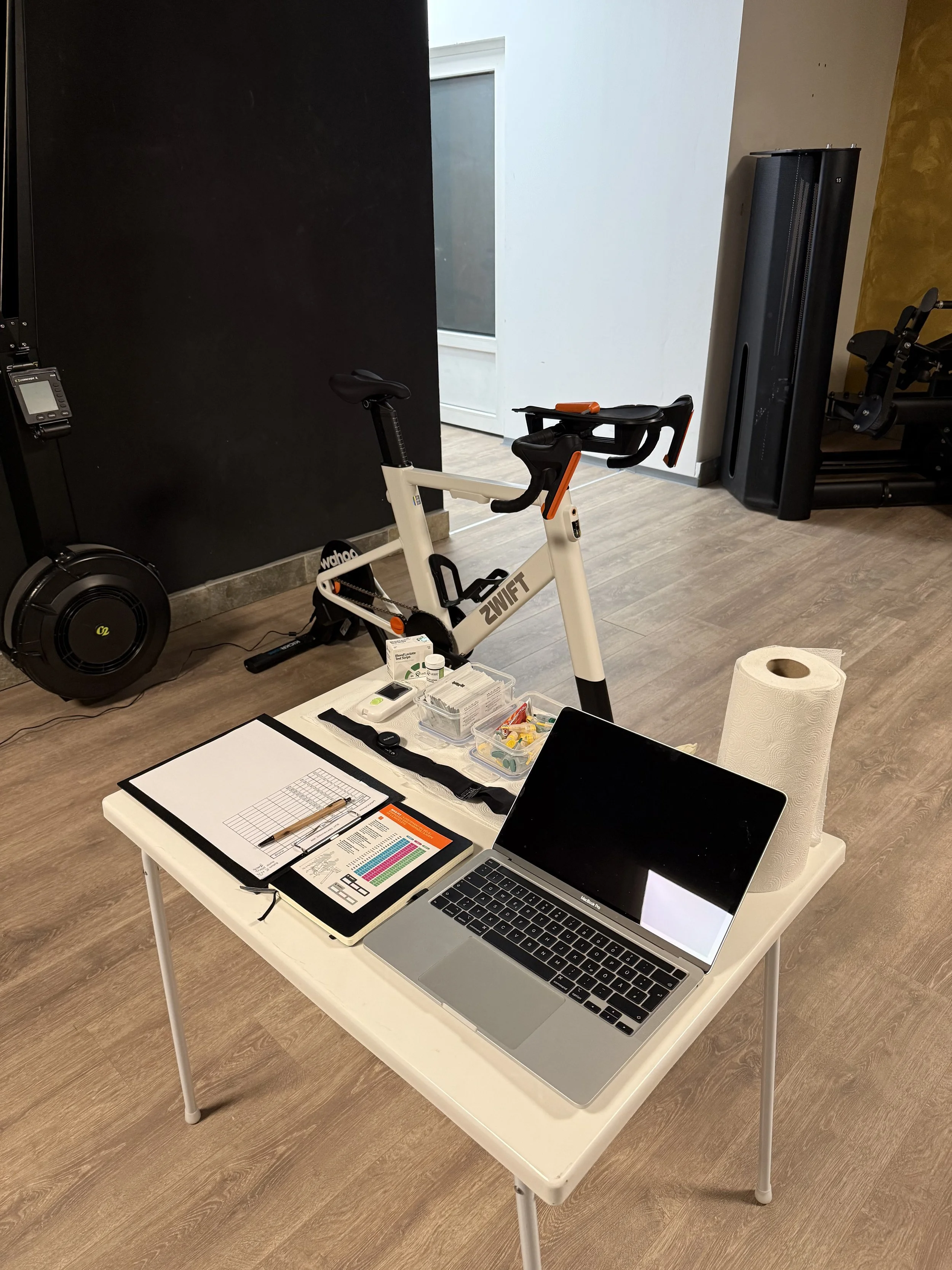 Ein Fitnessraum mit einem Hometrainer, einem Laptop, einem Notizblock, Medikamenten und einer Küchenrolle auf einem weißen Tisch.