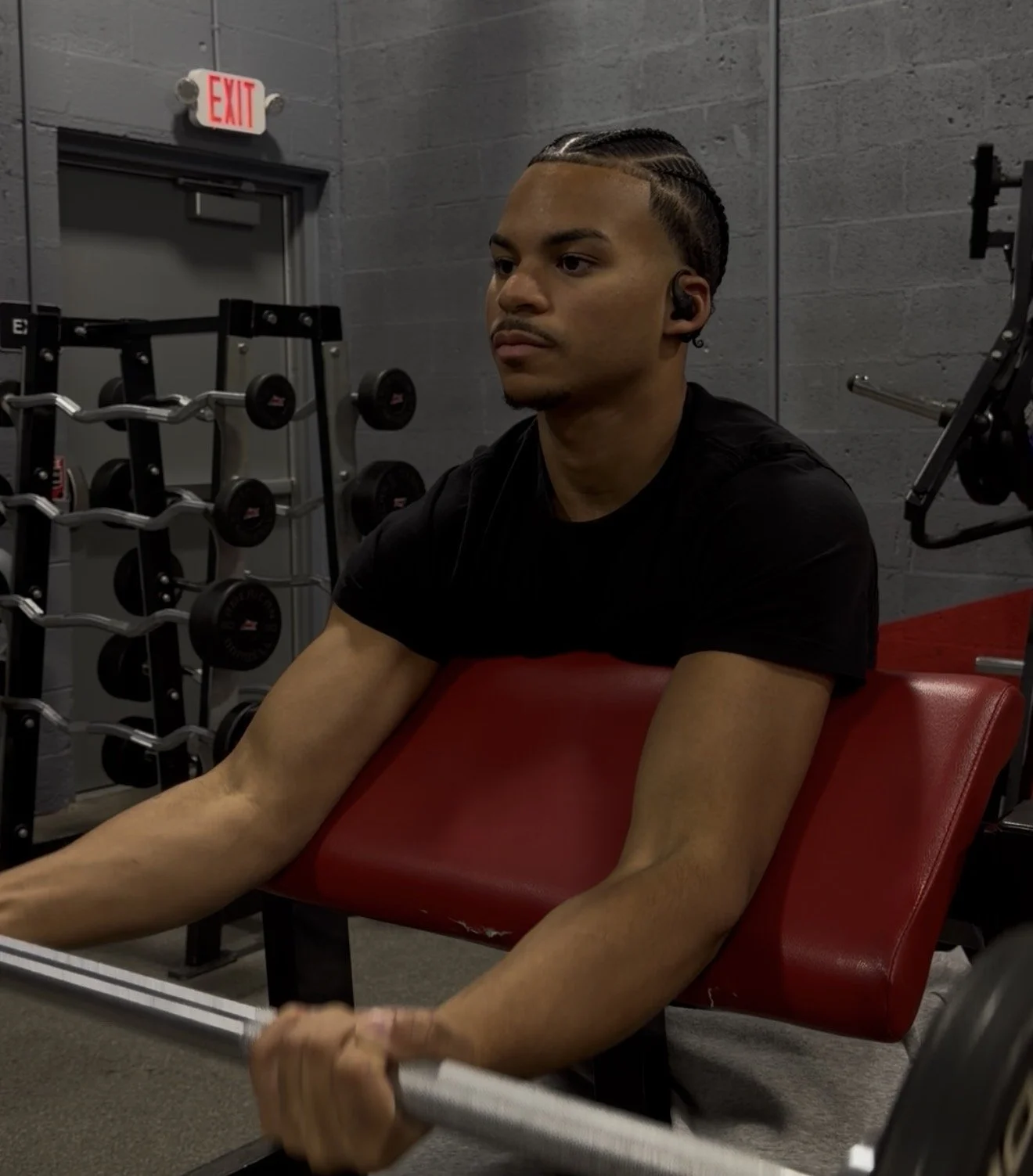 A young man with cornrow braids wearing a black shirt, sitting on a red workout bench at the gym, holding a barbell. Gym equipment and an exit sign are visible in the background.