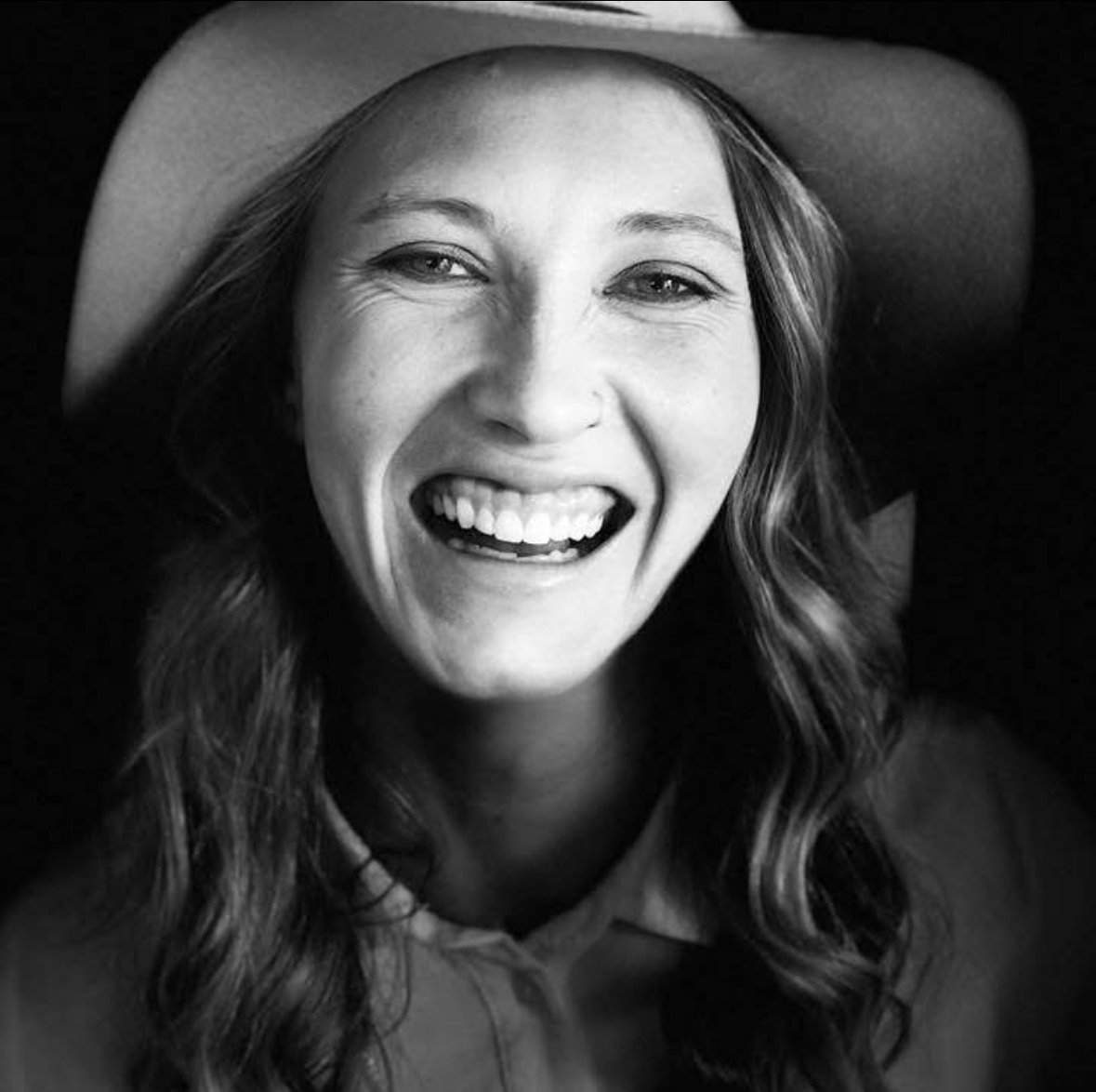 Black and white photo of a woman smiling widely, showing her teeth, with her head tilted back and her hair in loose waves.