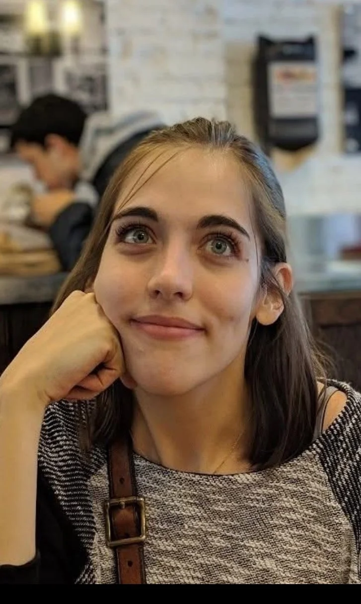 A young woman with blue eyes and long brown hair sitting at a table in a restaurant or café, resting her chin on her hand, looking slightly off to the side with a gentle smile.