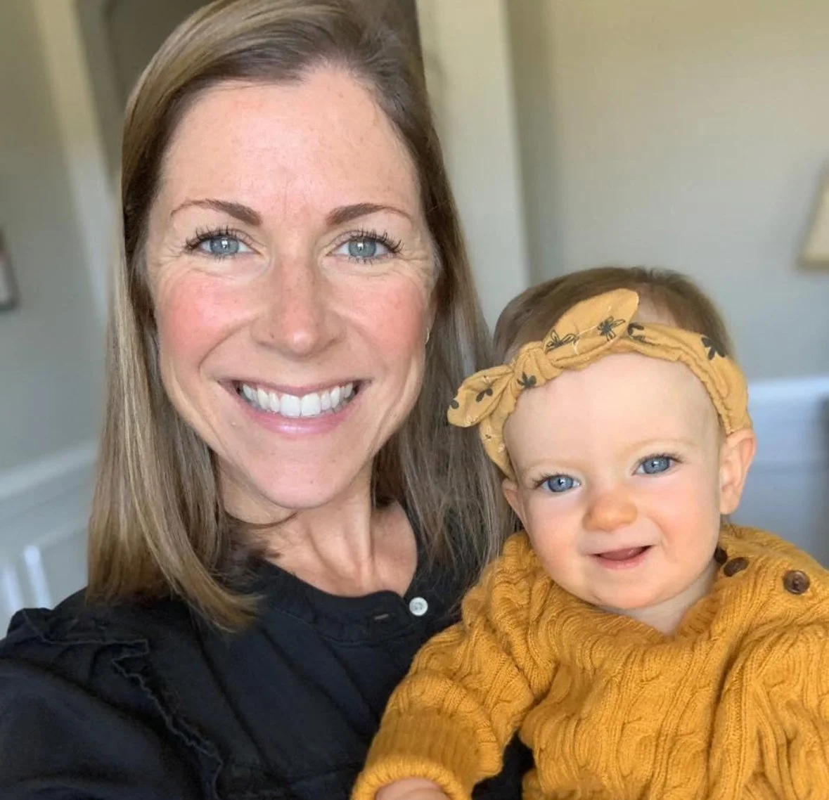 A woman with blonde hair and blue eyes smiling while holding a baby girl with blonde hair, blue eyes, and a yellow headband, dressed in a yellow sweater.