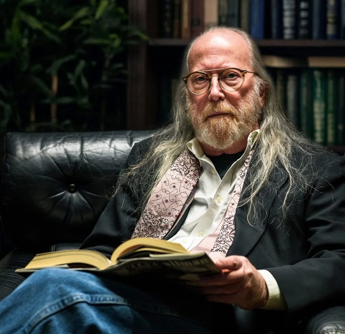 An older man with long gray hair, glasses, and a beard sitting on a black leather chair in a library or study, holding an open book.