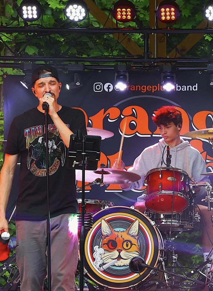 A band performing on an outdoor stage with green foliage in the background. A young man is singing into a microphone, and another young man is playing the drums. The drum set features a logo of a cat with one eye, smoking a cigarette. The backdrop displays social media icons and the band name 'Orange Plus Band'.