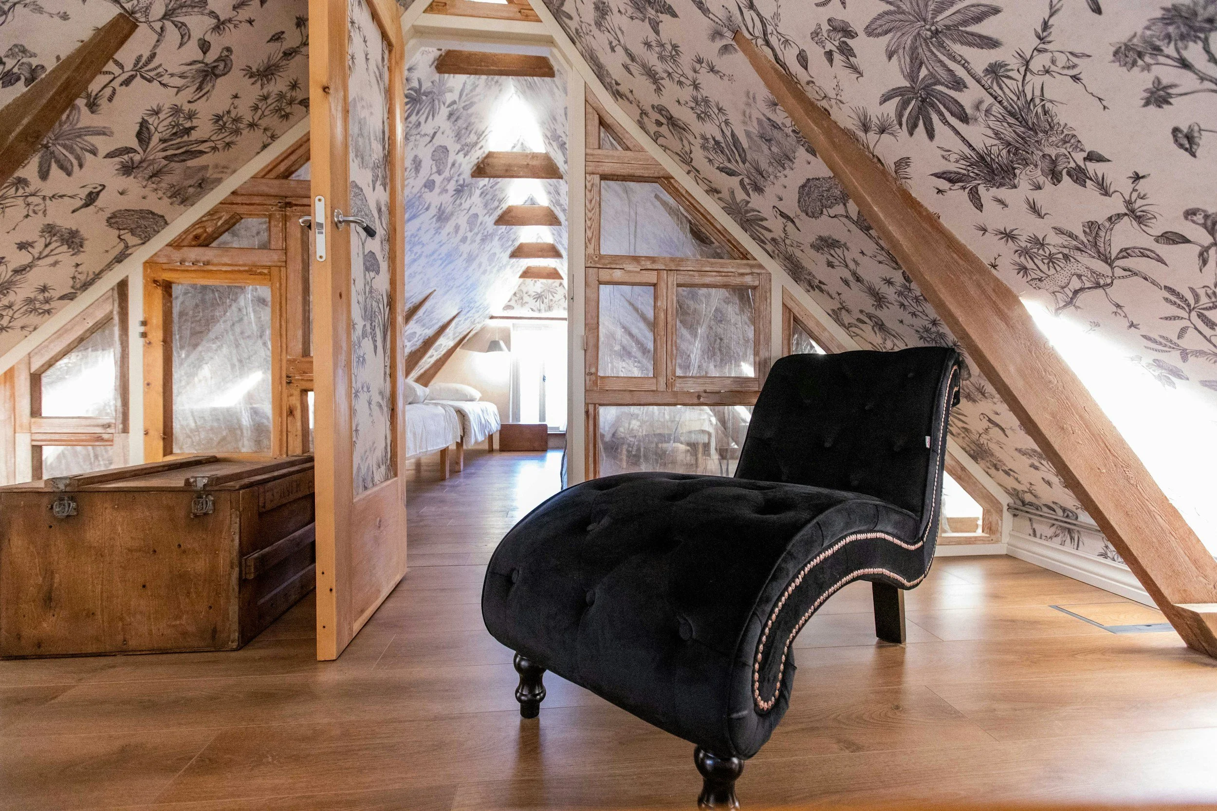 A cozy attic room with sloped walls and ceiling covered in floral wallpaper, wooden beams, a black velvet chaise lounge, a wooden trunk, and an open door leading to a bedroom with a bed and window with sunlight.