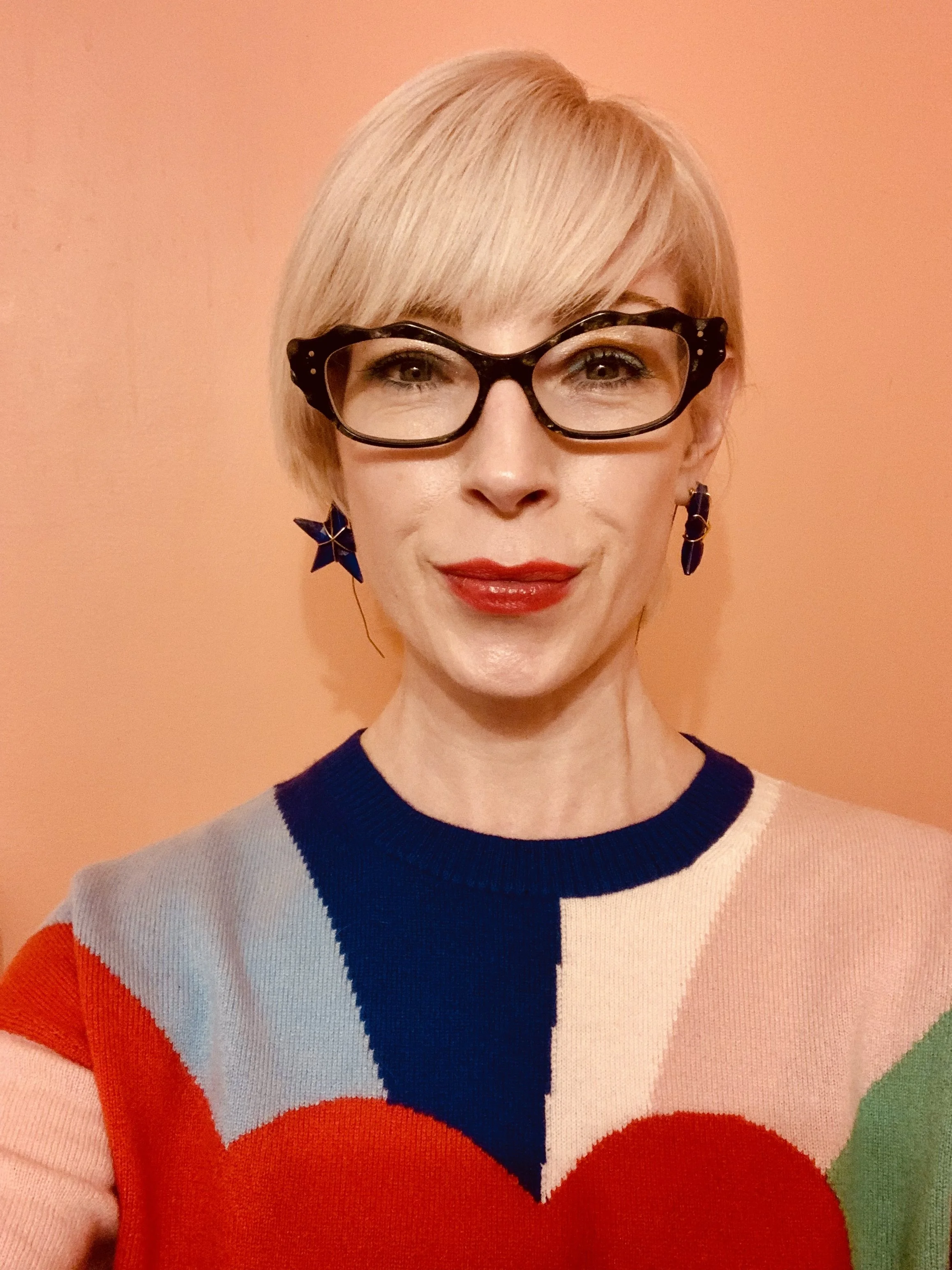 A woman with short blond hair, wearing black glasses, red lipstick, and star-shaped earrings, smiling against a peach background, dressed in a colorful sweater with geometric patterns in red, blue, pink, and green.