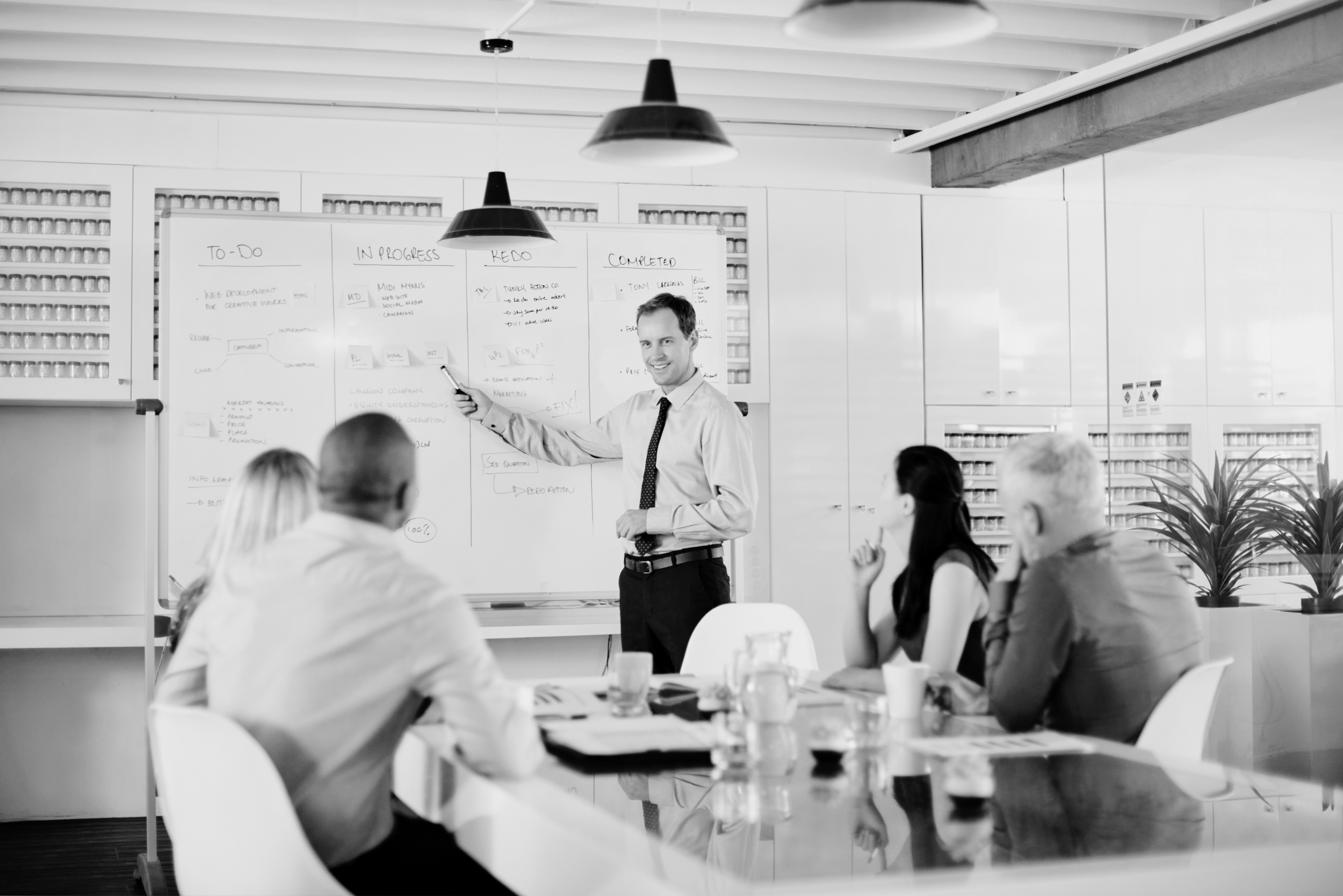 A man in a shirt and tie giving a presentation to four people sitting at a table in a conference room. The man is pointing at a whiteboard filled with handwritten notes.