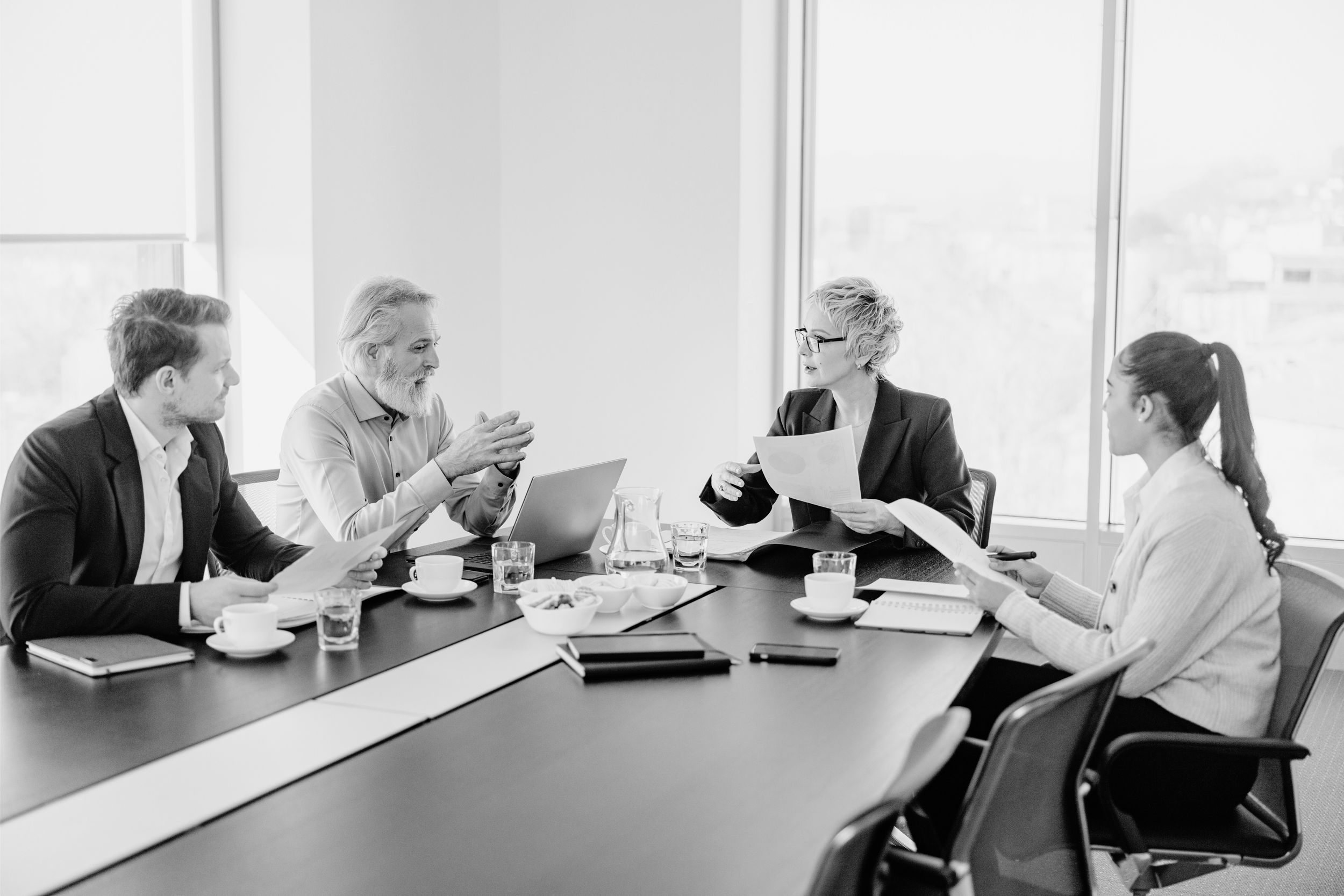 Four people in business attire having a discussion in a modern conference room with large windows.