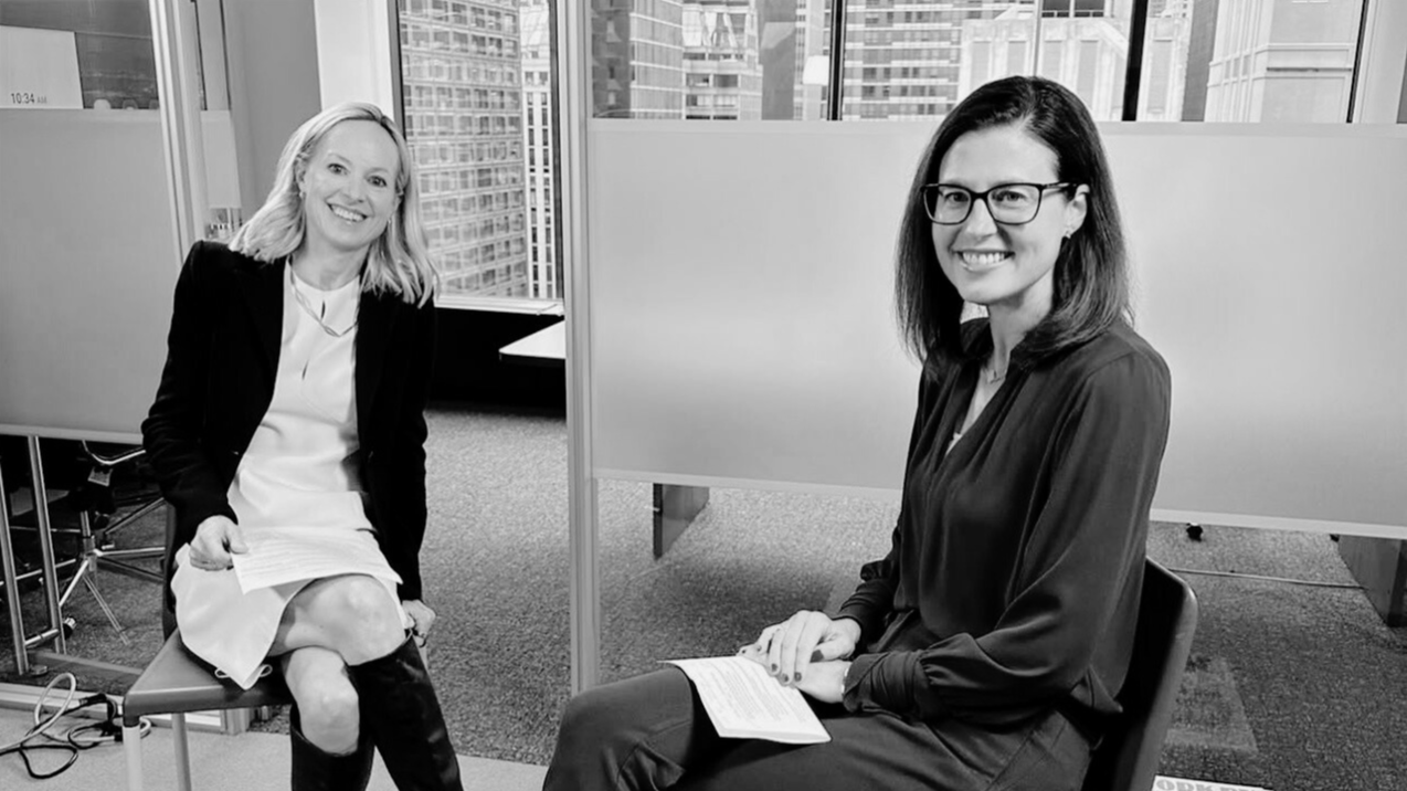 Two women sitting on chairs in an office with large city buildings visible through windows, smiling at the camera.