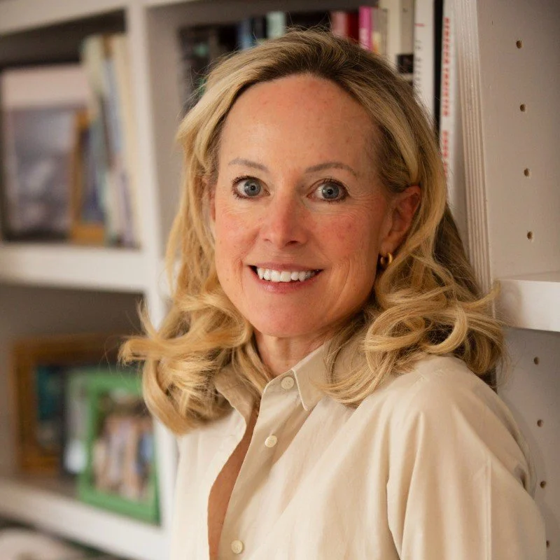 A woman with blonde curly hair, wearing a cream-colored blouse and small hoop earrings, smiling and standing next to a bookshelf filled with various books.