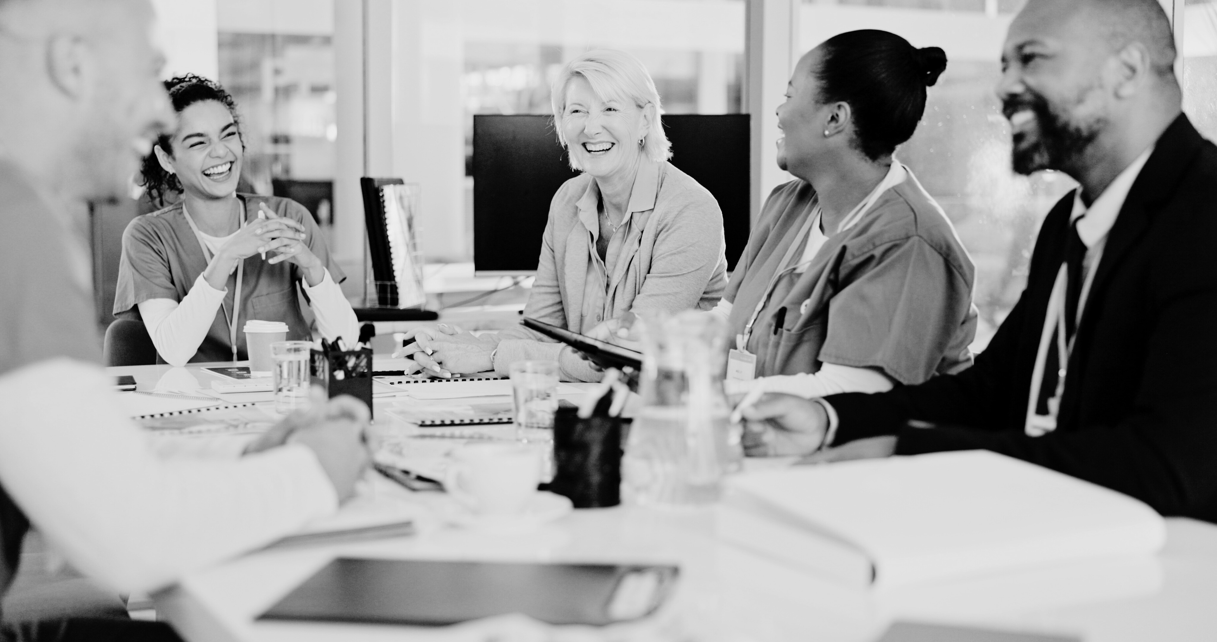 A diverse group of healthcare professionals, including nurses and doctors, are sitting around a table in a meeting room, laughing and sharing a moment of joy.