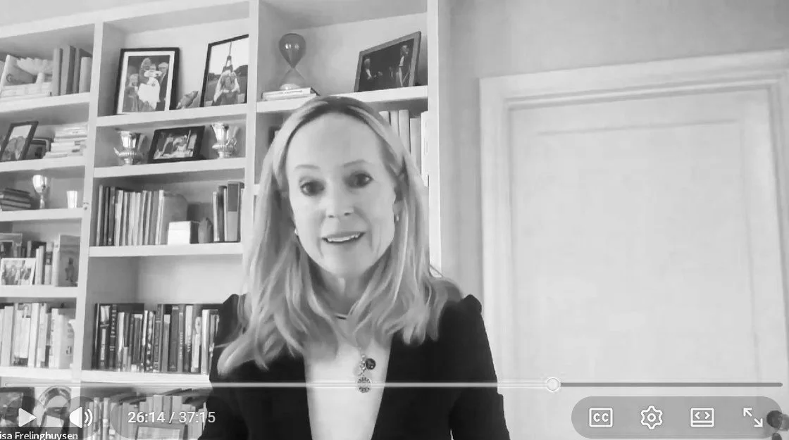 A woman with blonde hair smiling at the camera in a room with a bookshelf filled with books and framed photos on the wall behind her.