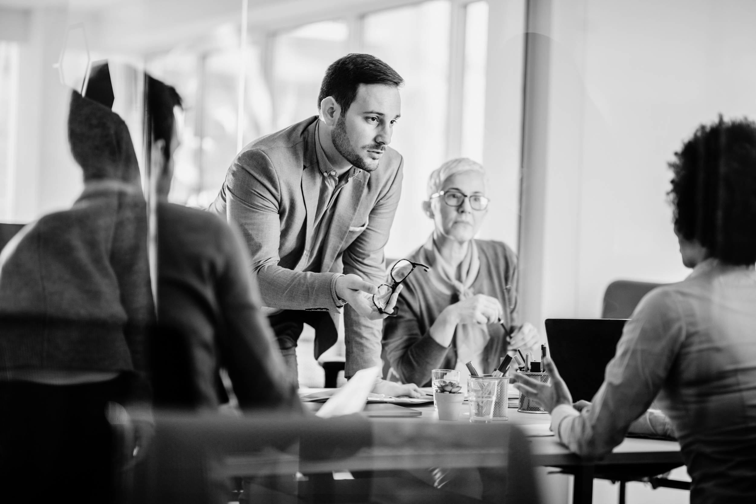 Business meeting with five people in a conference room, one man standing and talking, holding glasses, others listening, some with papers and glasses, in black and white.