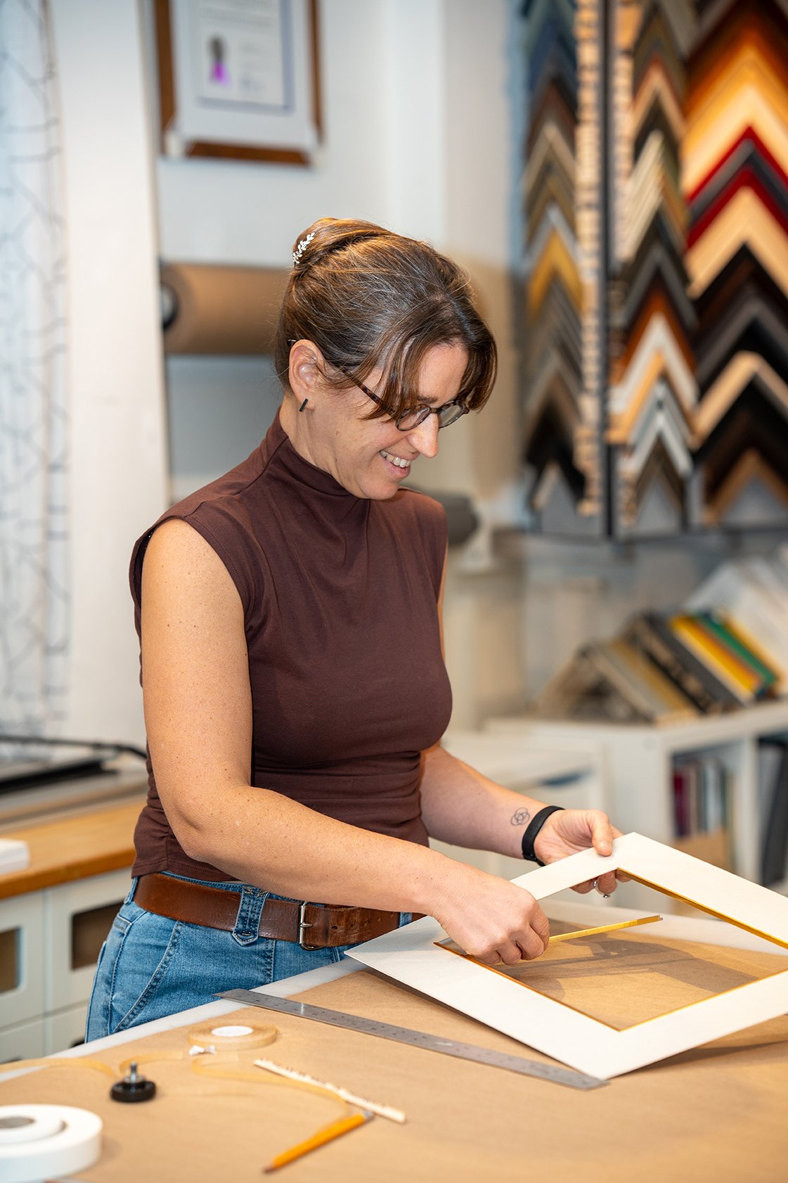 A woman in a brown sleeveless top and glasses is smiling while examining a picture frame in a framing shop. There are framing supplies on the table and samples of framing styles and colors on the wall behind her.