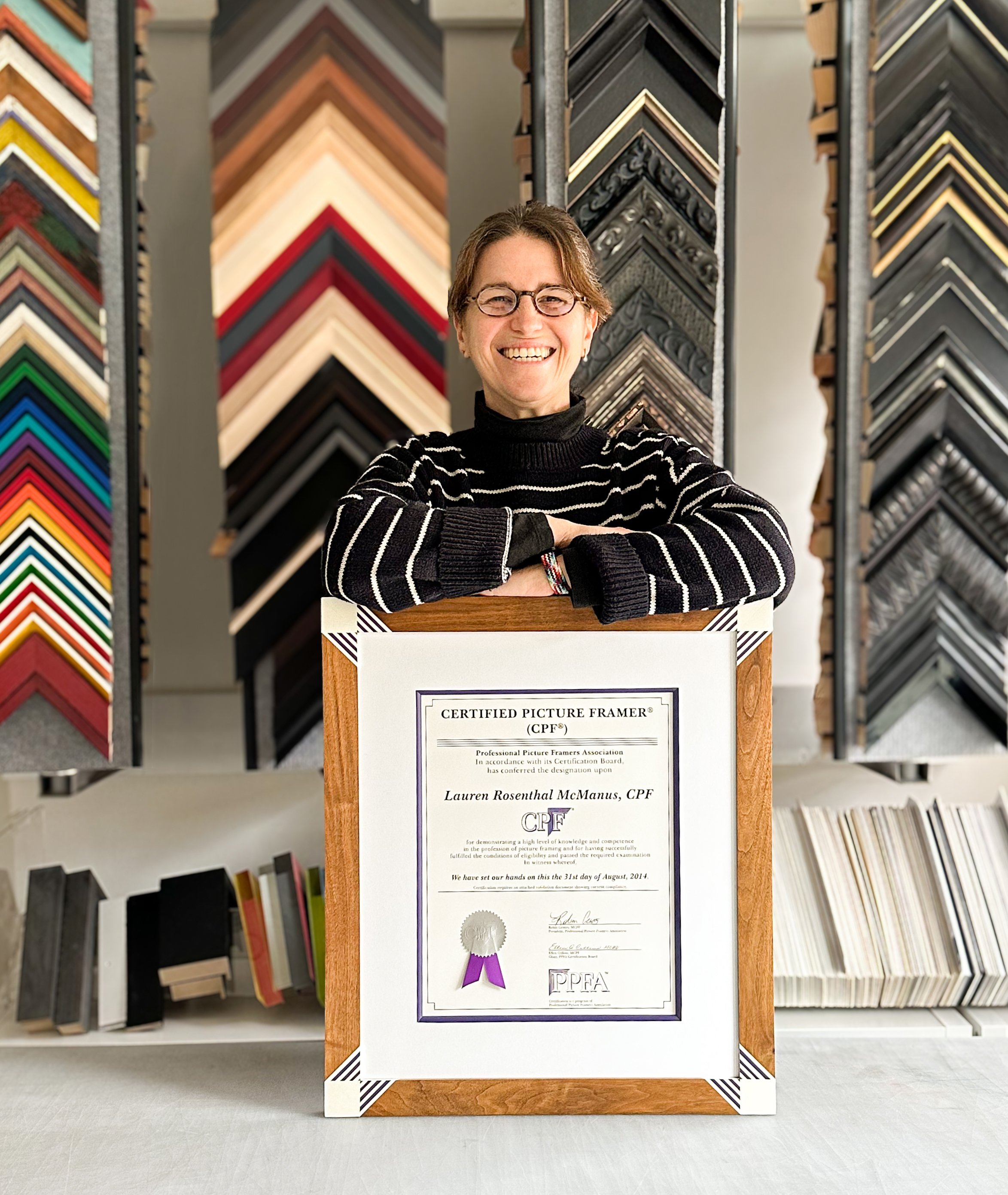 A smiling woman with glasses and a black and white striped sweater displays a framed certificate in a store with various picture frame moldings on display behind her.