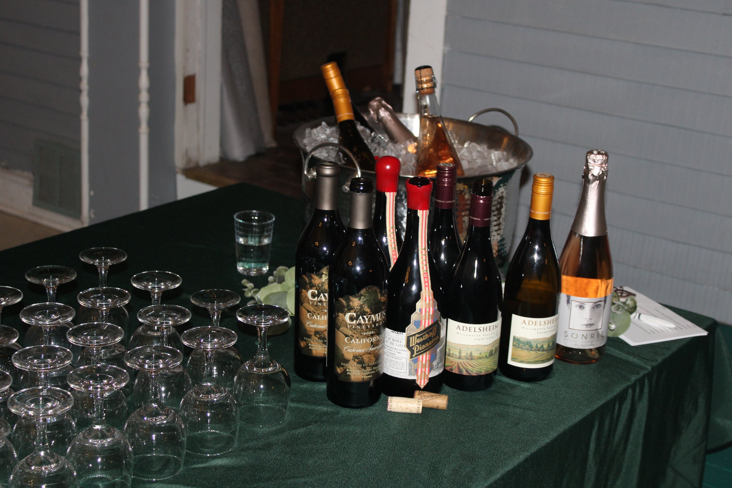 A table with several bottles of wine, some with red caps and one with a white and black label. There are many empty wine glasses on the table, which is covered with a dark green tablecloth. In the background, there is a metal ice bucket filled with i