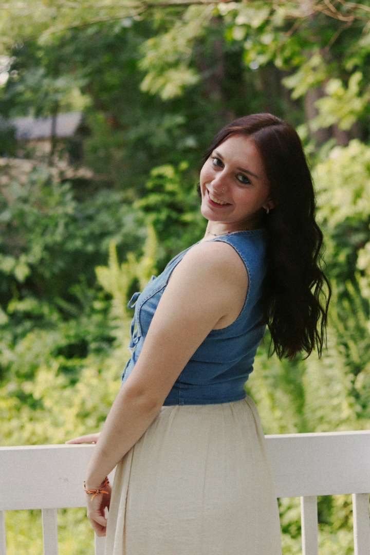 A woman with long dark hair smiling and leaning on a white railing outdoors, with green trees in the background.