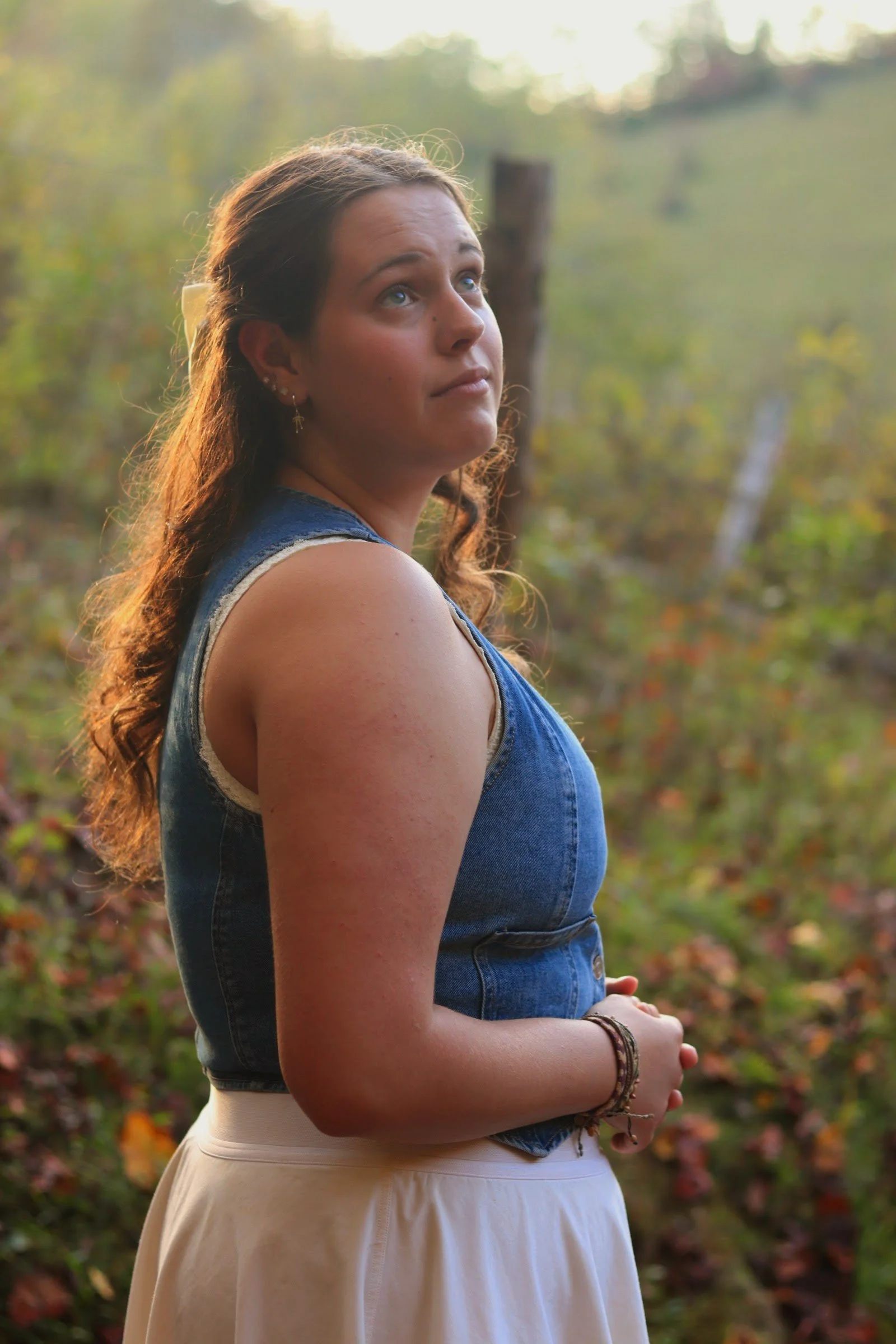 A young woman standing outdoors in a natural setting with trees and foliage in the background. She has long, wavy brown hair, light skin, and is wearing a sleeveless denim top and a beige skirt. She is looking slightly upward and to the left, with her arms gently crossed and wearing bracelets and earrings.
