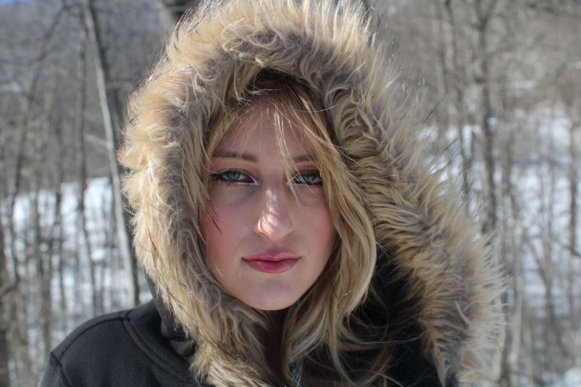 A woman outdoors in winter wearing a black jacket with a fur-lined hood. Snowy trees are in the background, and she has blond hair, blue eyes, and natural makeup.