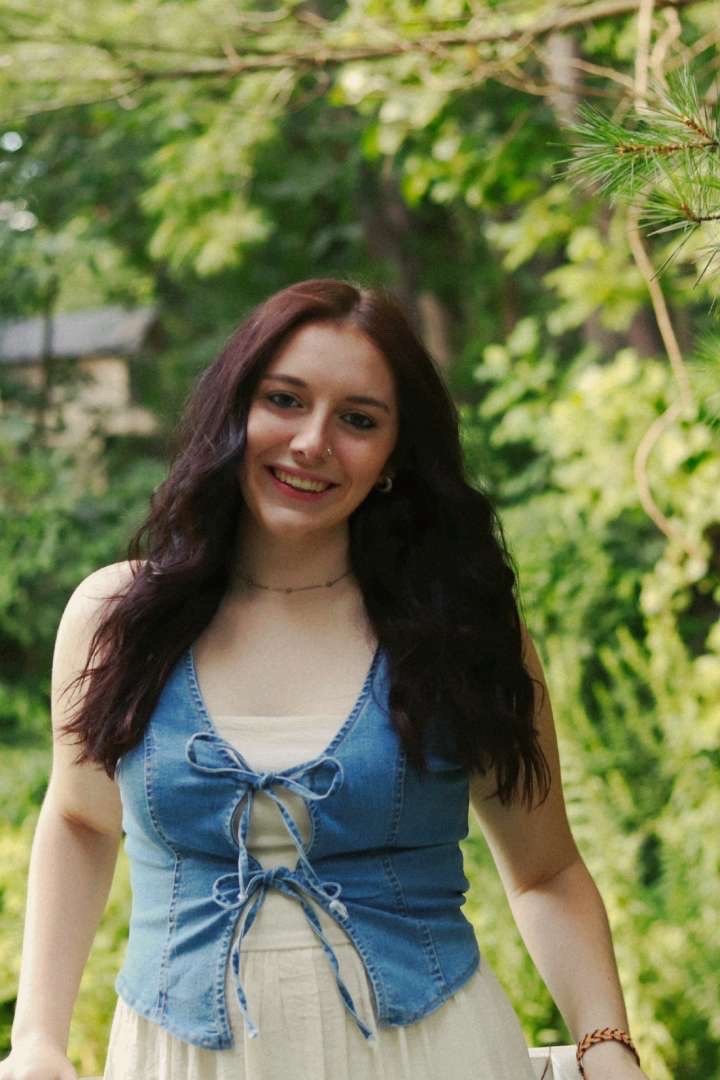 A young woman with long, wavy dark hair smiling outdoors in a lush green garden, wearing a denim sleeveless top with bow details and a light-colored skirt.