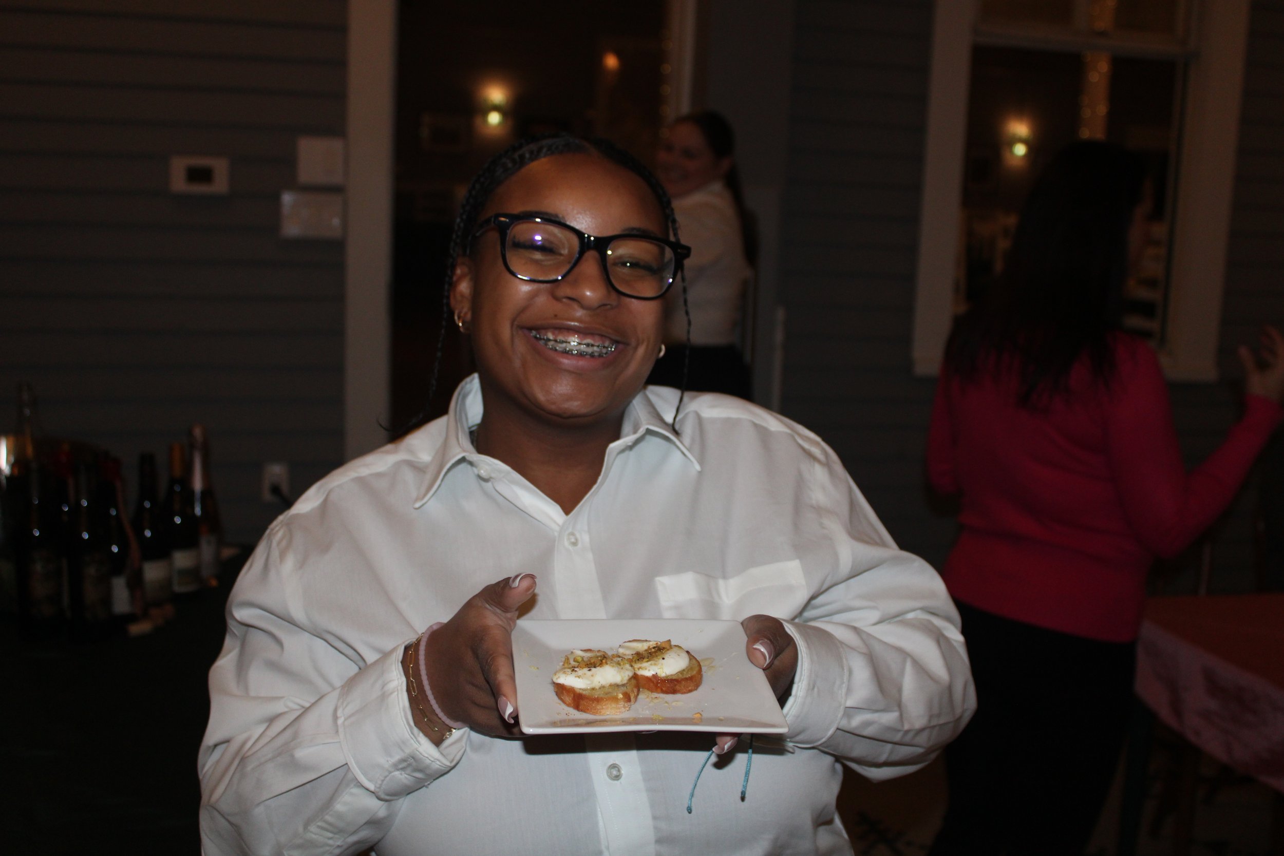 A woman wearing glasses and a white shirt smiling and holding a plate of bruschetta at a social gathering.
