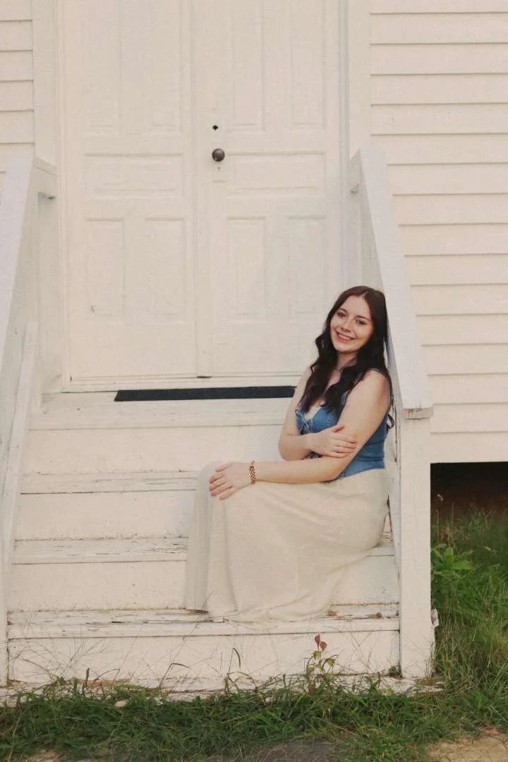 A young woman with long dark hair, wearing a blue top and a long beige skirt, sitting on white wooden stairs outside a house, smiling at the camera.
