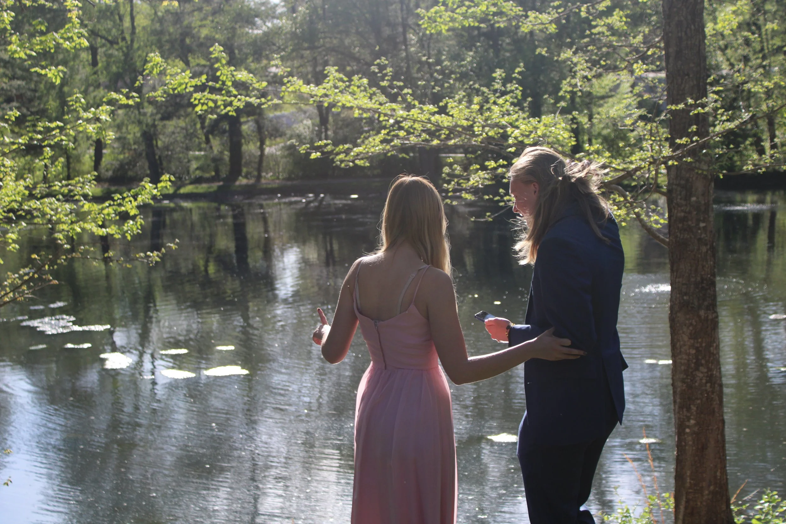 A woman in a pink dress and a man in a dark jacket are standing by a lake in a green wooded area, engaged in conversation while looking at a phone.