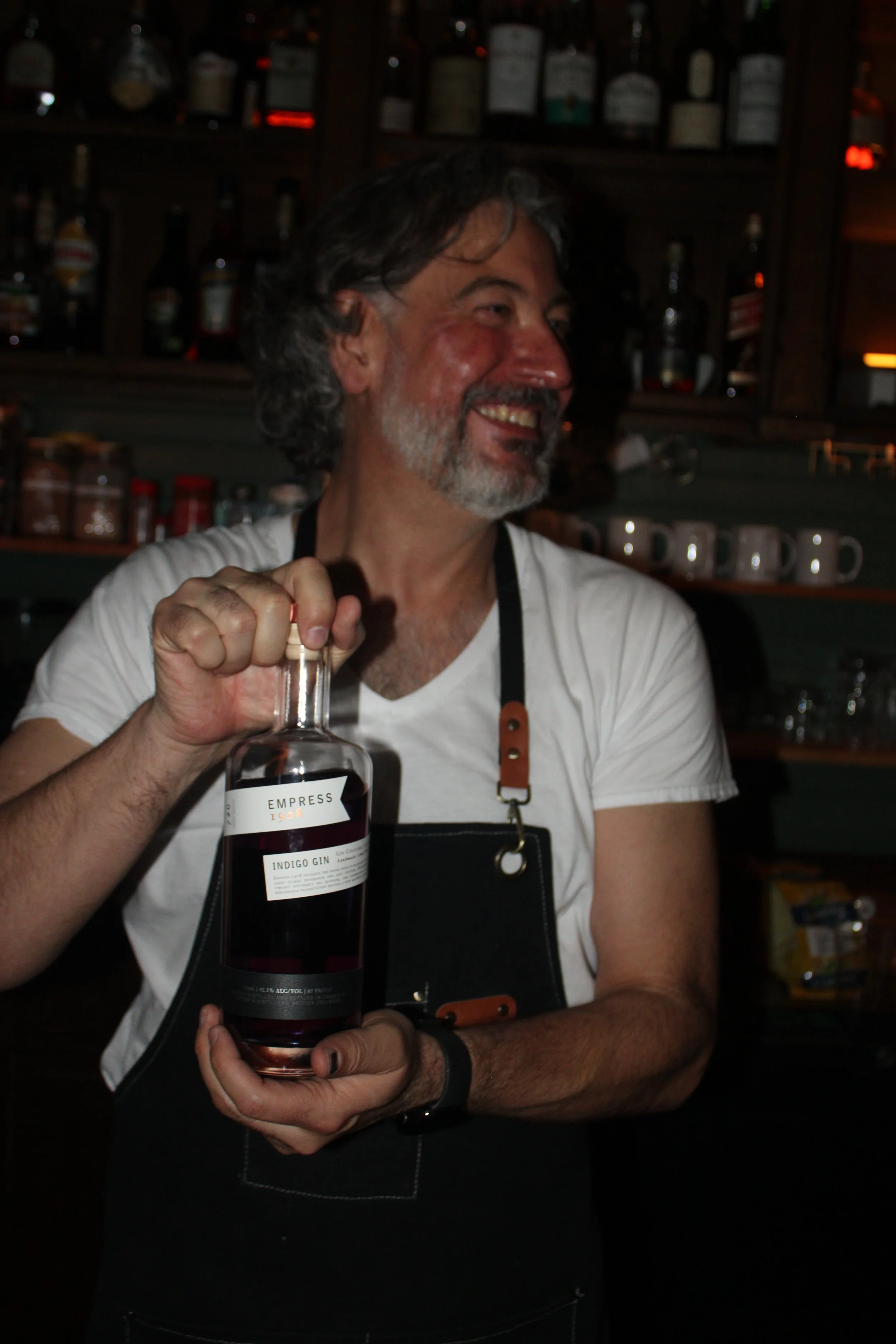 A man with gray hair and a beard smiling as he holds a bottle of gin in a bar setting with shelves of bottles in the background.