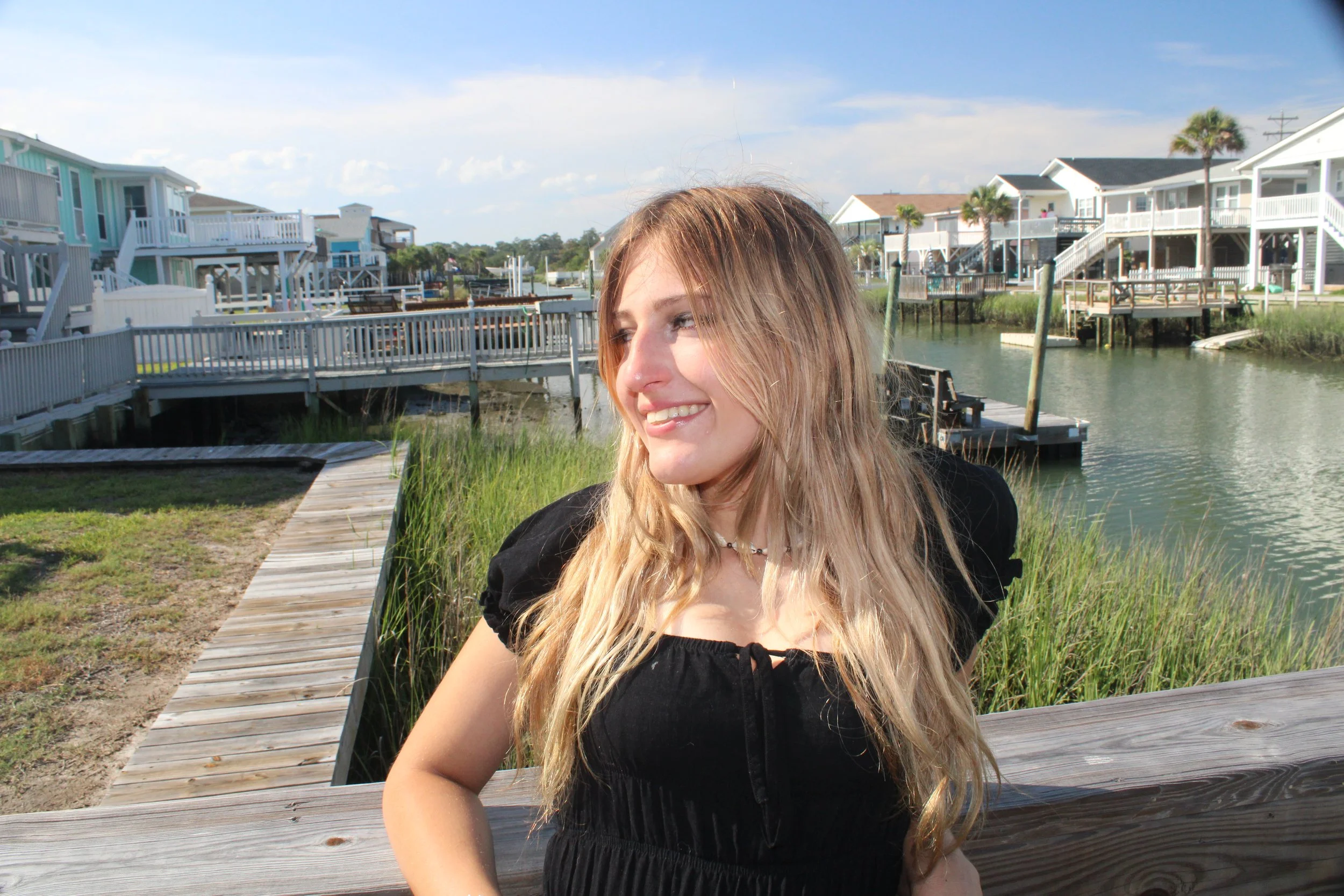 A woman with long blonde hair smiling outdoors near a waterway with colorful houses on stilts and a blue sky in the background.