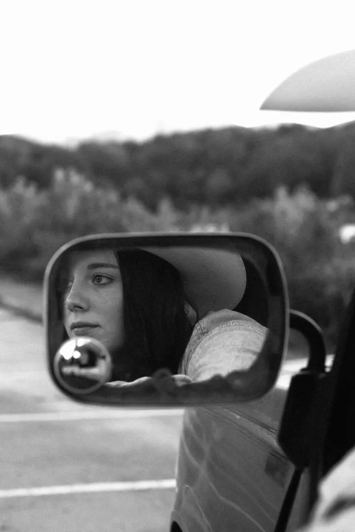 A woman looking out from the side mirror of a vehicle, with a landscape of trees and hills in the background, in black and white.
