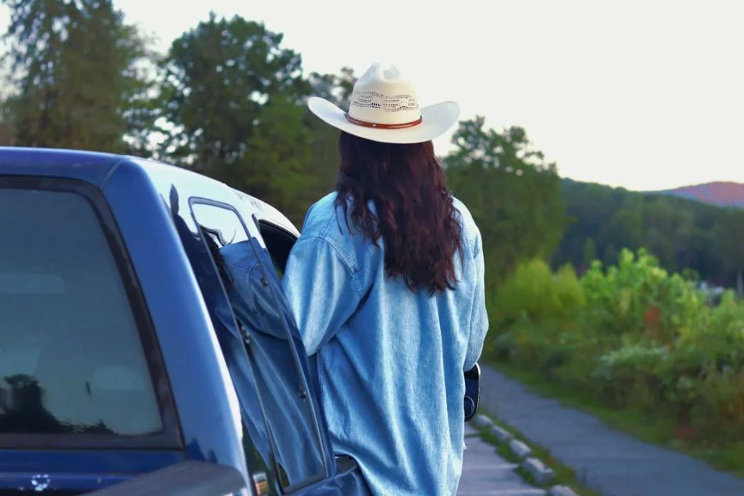A woman with long curly hair wearing a tan cowboy hat and a light blue denim jacket standing outside near a blue vehicle on a rural road, with trees and hills in the background.