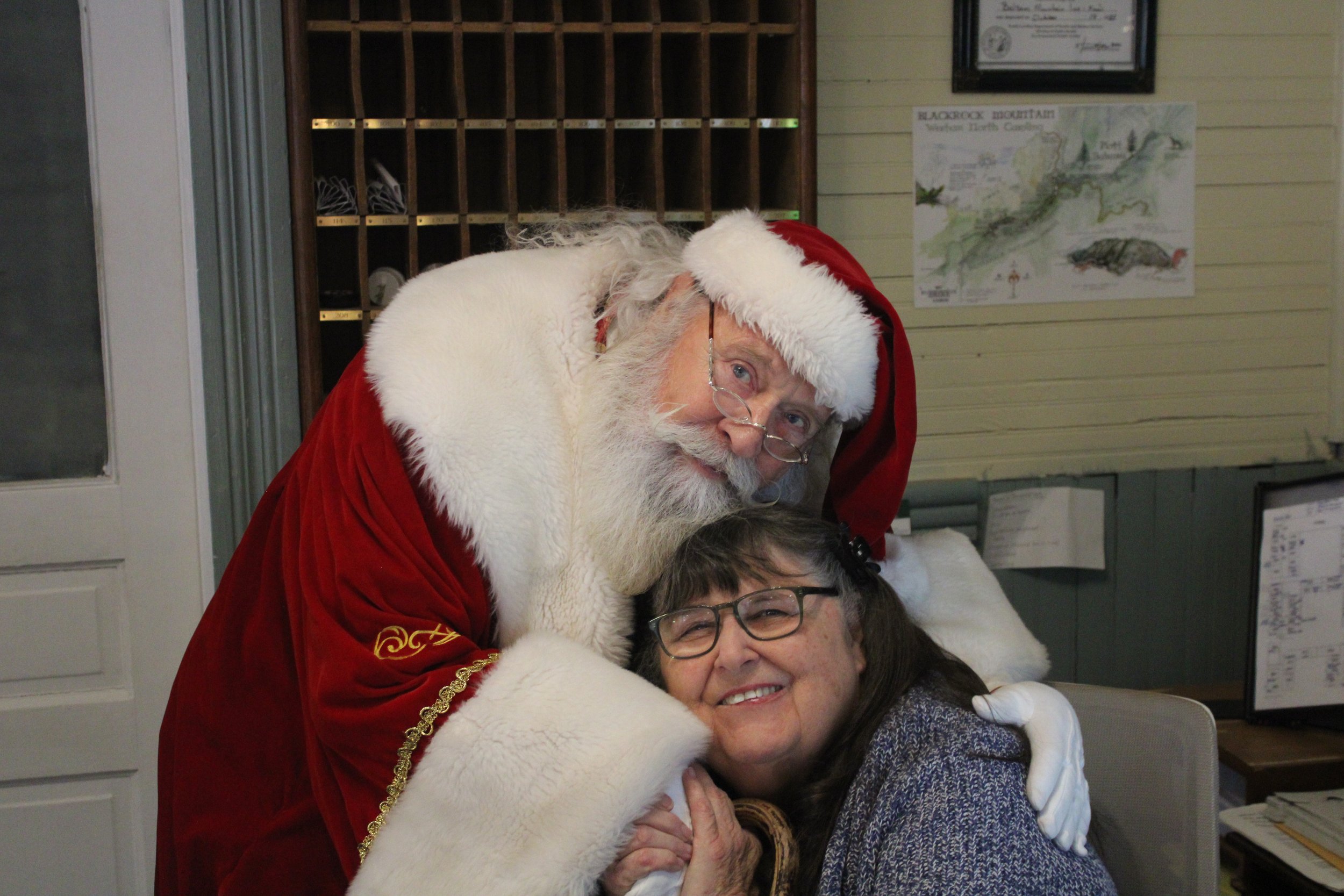 Santa Claus in a red and white suit leaning over one smiling women, one of whom is wearing glasses and a gray sweater. They are indoors with a wooden mail organizer and a map on the wall in the background.