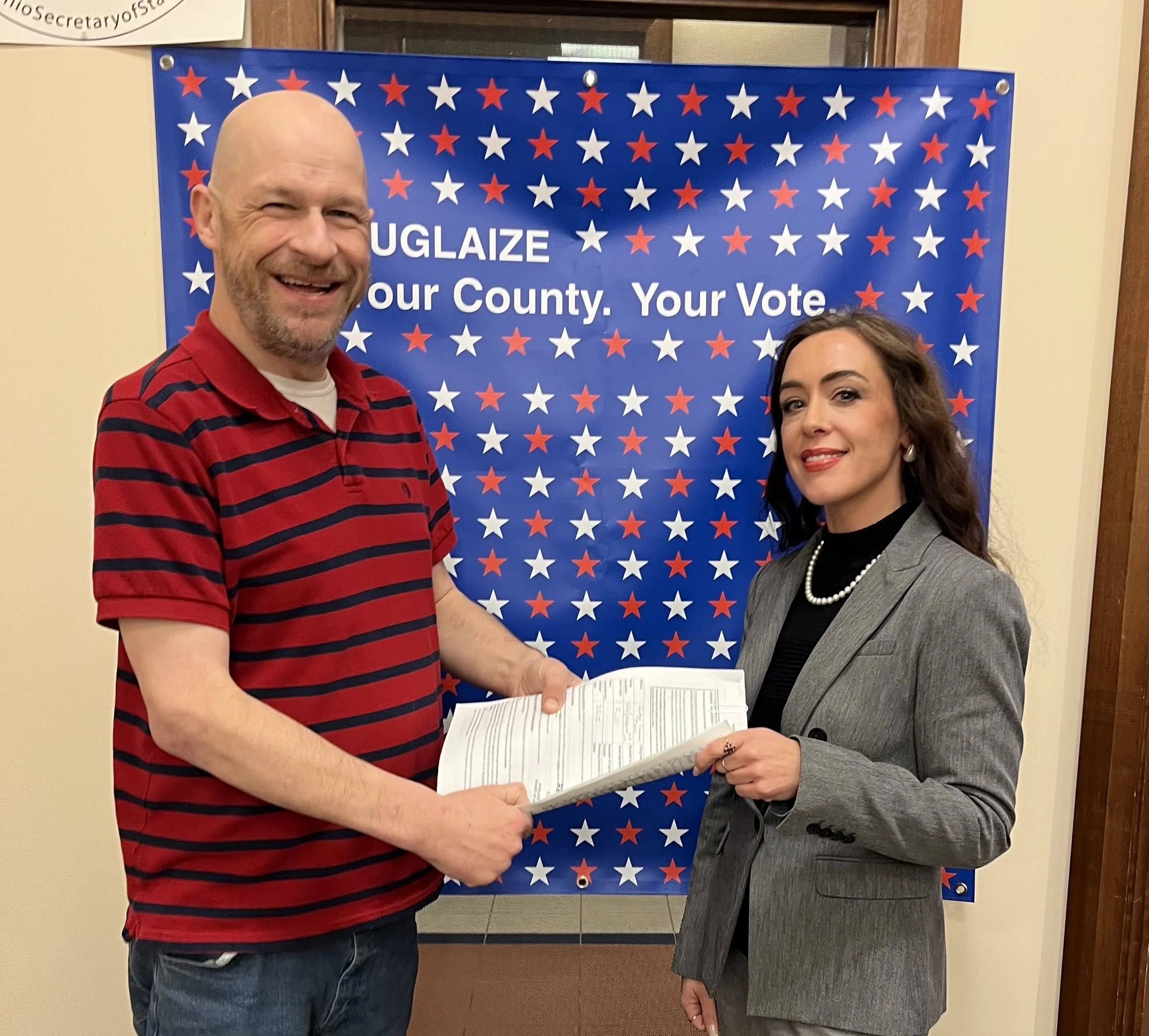 A man and a woman stand together holding a document in front of a patriotic themed background with red, white, and blue stars. The man is wearing a red and navy striped polo shirt, and the woman is wearing a gray blazer over a black top with a pearl 