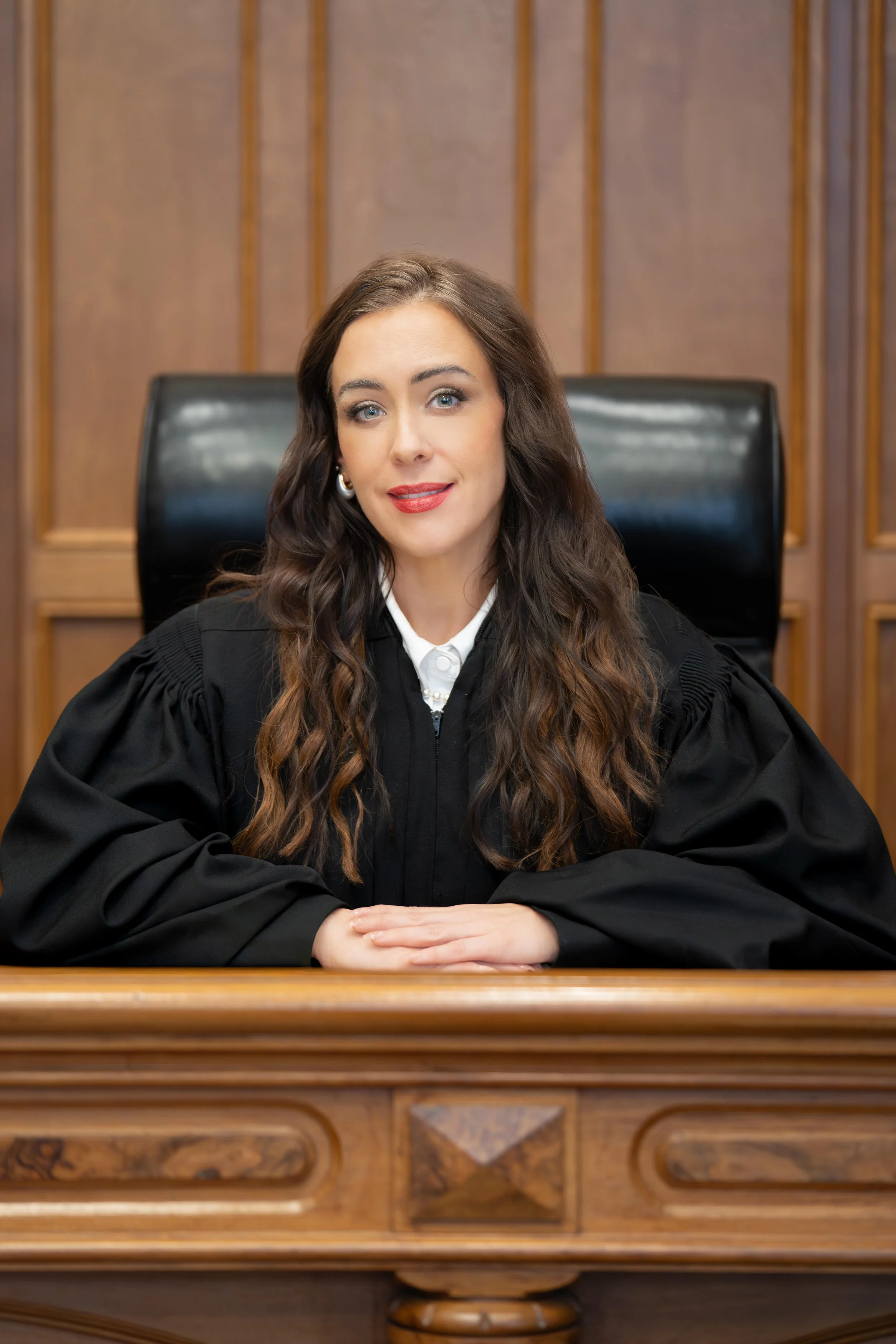 A female judge sitting at a wooden bench in a courtroom, wearing a black robe and looking at the camera.