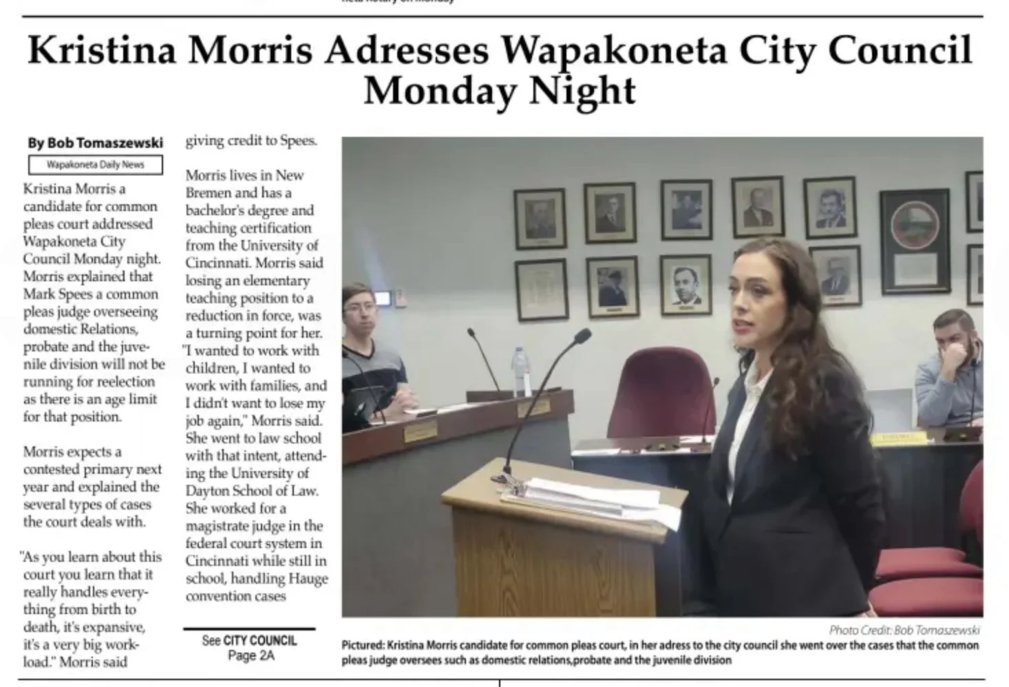 Kristina Morris, a candidate for common pleas court, speaking at a city council meeting, with two other people seated behind her in a room with framed photographs on the wall.