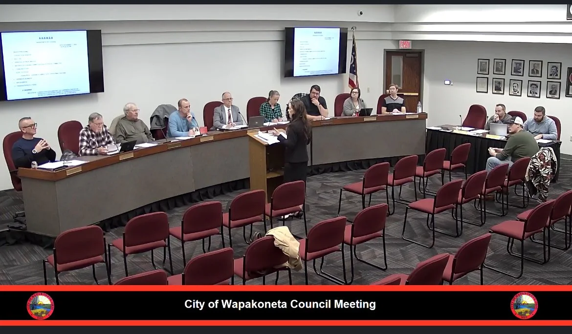 City of Wapakoneta Council Meeting room with multiple people seated at a long table, a woman speaking at a podium, and empty chairs facing the panel. Large screens display documents, and framed pictures are on the wall.