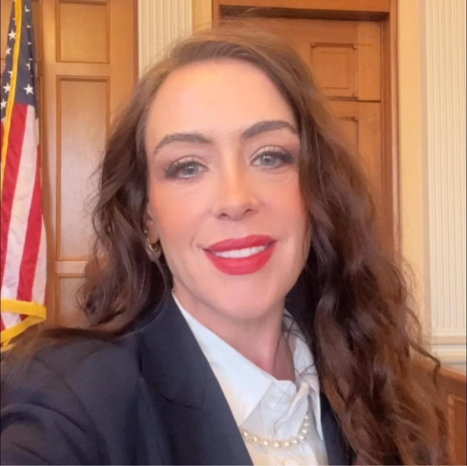 A woman with wavy brown hair, red lipstick, and blue eyes, wearing a dark blazer, white shirt, and a pearl necklace, in a room with wooden paneling and an American flag in the background.