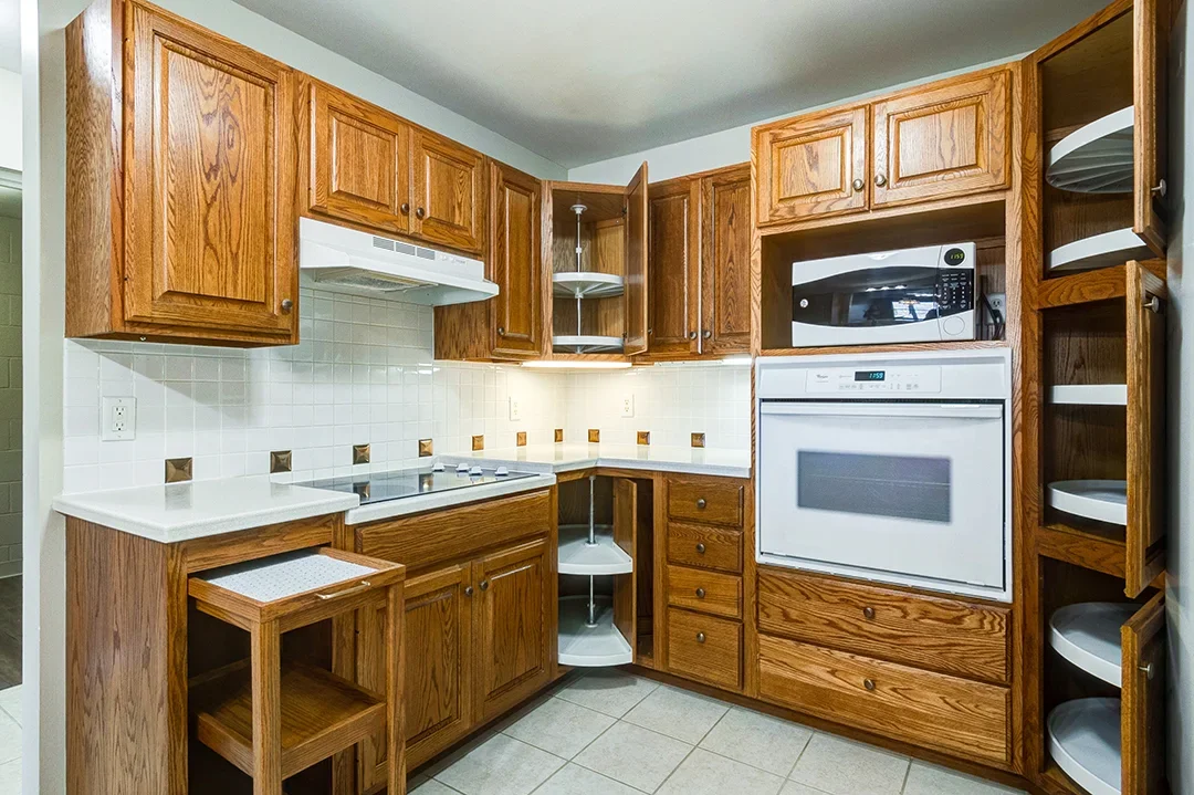 Corner kitchen with oak cabinets, white countertops, built-in microwave, oven, and open shelves, tiled backsplash, and tiled floor.