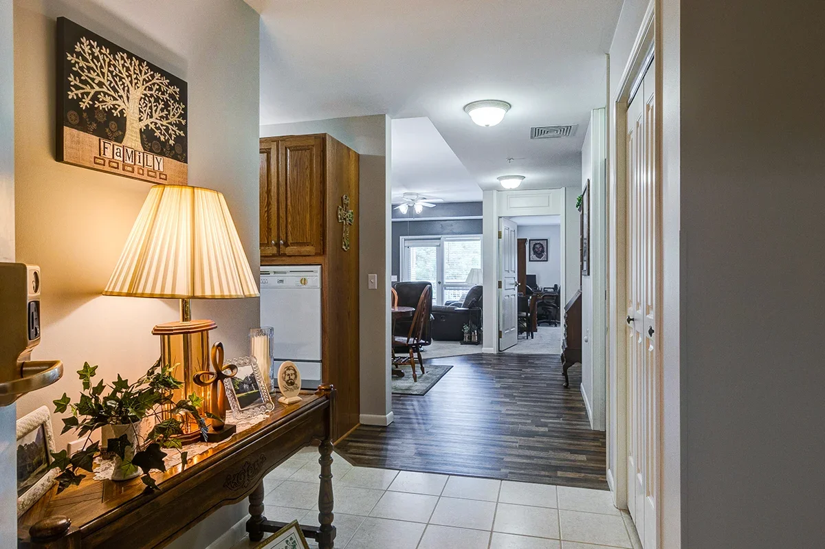 Entrance hallway with a wooden console table, lamp, picture frames, and decorative leaves, leading into a dining and living area with wooden furniture, a ceiling fan, and large windows.