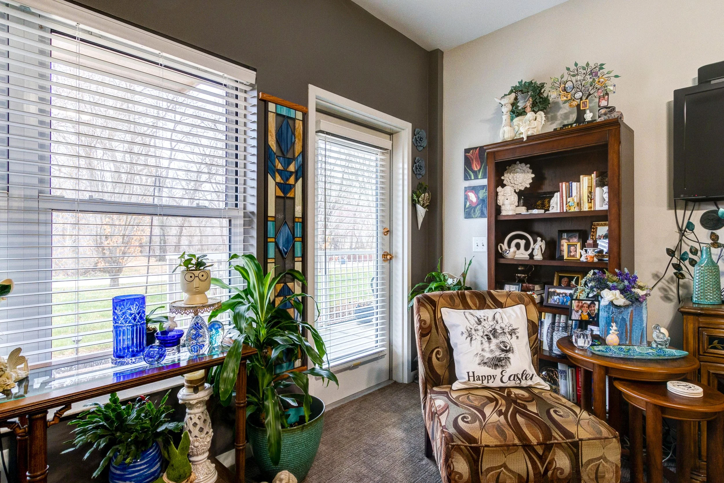 Living room with large windows, a door, and decorating items, including plants, glassware, paintings, and a chair with a "Happy Easter" pillow.