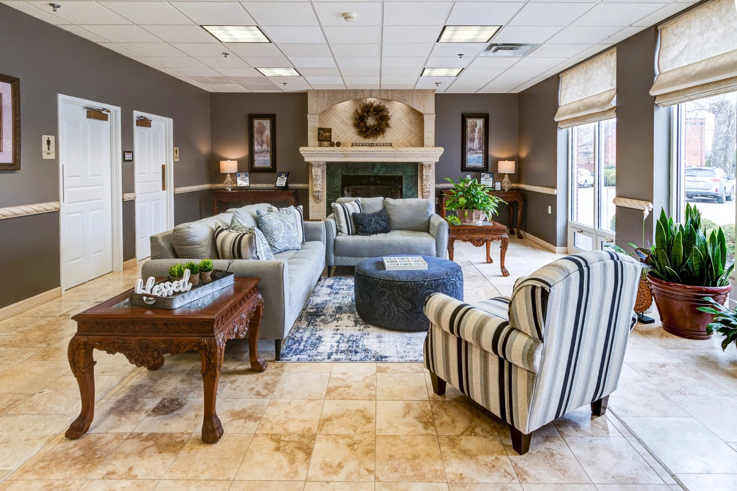 A spacious hotel lobby with a cozy seating area, including a gray sofa, striped armchair, and a round black Ottoman. There are potted plants, decorative pillows, side tables with lamps, and a central fireplace with a wreath above it. Large windows on