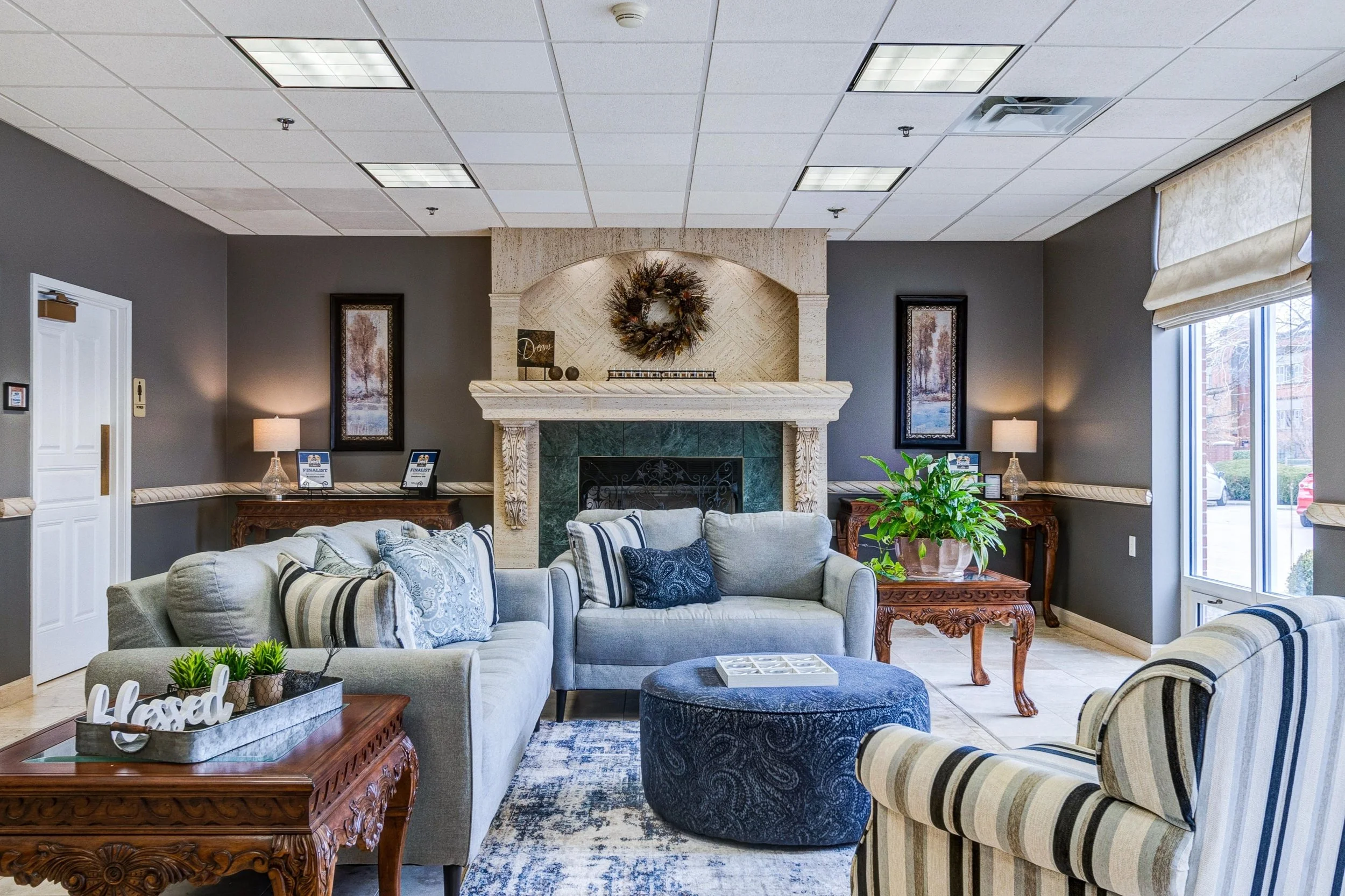 A cozy hotel lobby with gray walls, a fireplace with a decorative wreath, and furnished with sofas, armchairs, and a ottoman. There are side tables with lamps, framed artwork, and a large window allowing natural light.